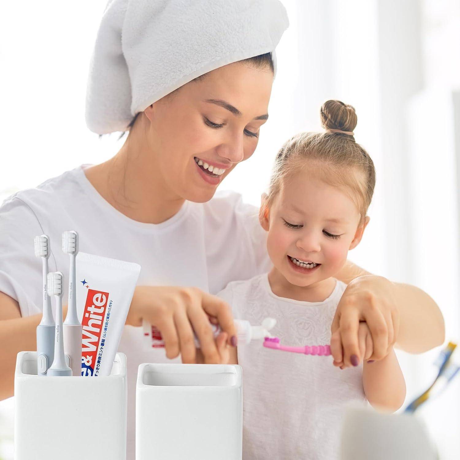 Lächelnde Mutter und kleine Tochter putzen sich gemeinsam die Zähne neben einem Behälter mit Zahnbürsten und einer Tube „Best White