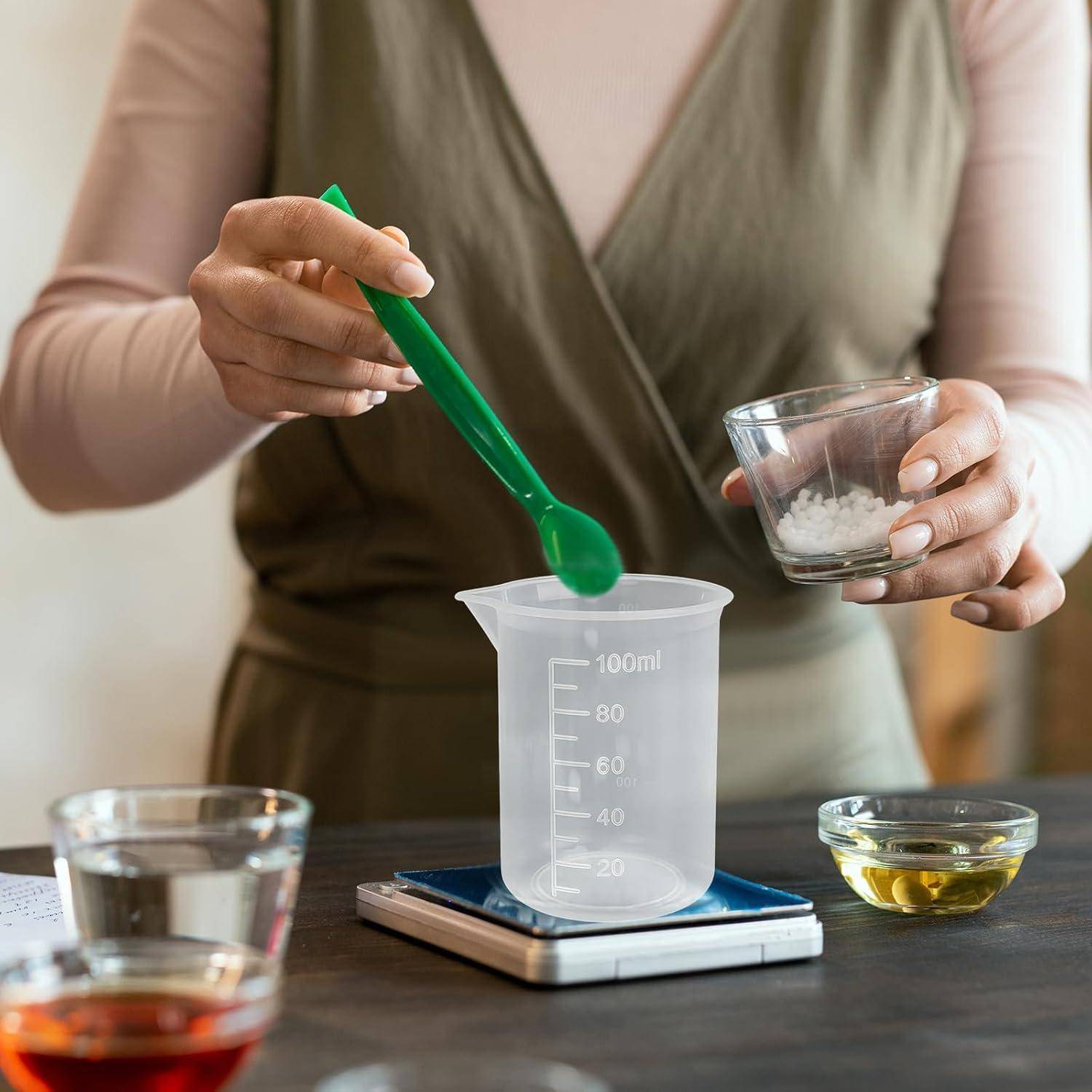 Eine Person misst Zutaten in einen Kunststoffbecher auf einer digitalen Waage ab, wobei sie einen grünen Löffel und eine kleine Schüssel im Küchenarbeitsbereich hält.