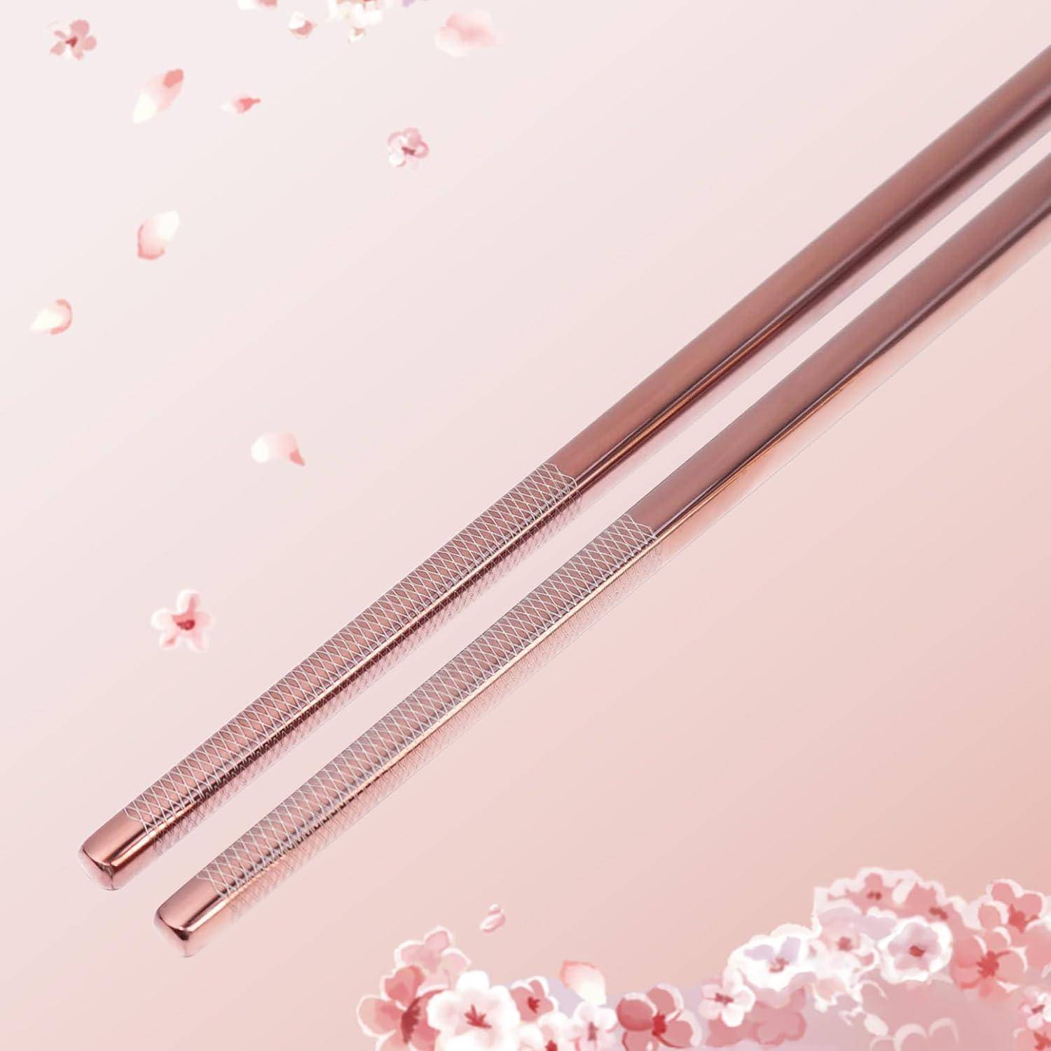 Paar rosa rosengoldfarbene Metallstäbchen auf zartem rosa Hintergrund mit verstreuten Kirschblütenblattpetalen.