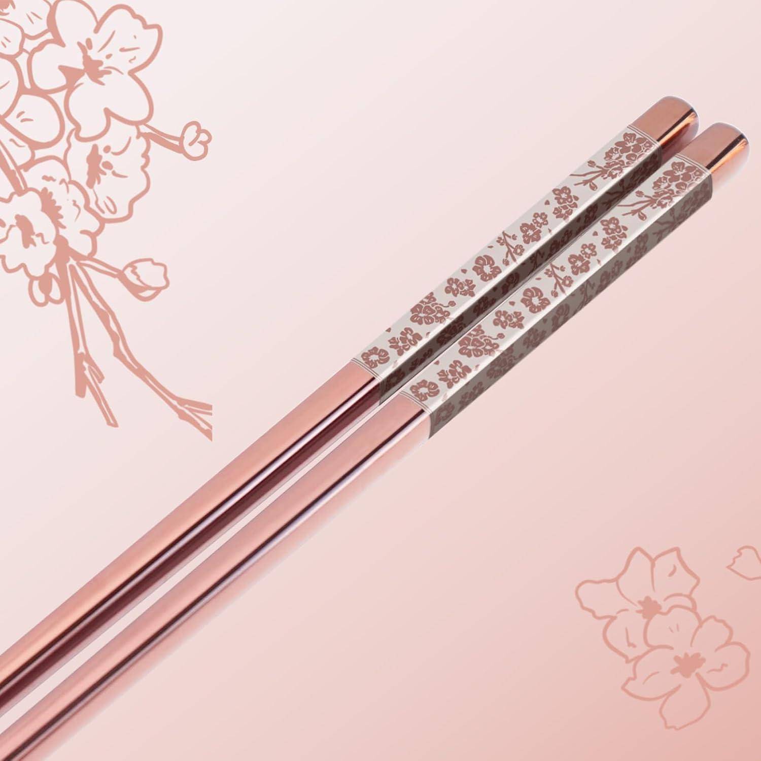 Zwei blütengemusterte rosegoldfarbene Essstäbchen auf hellrosanem Hintergrund mit Kirschblütenillustrationen.