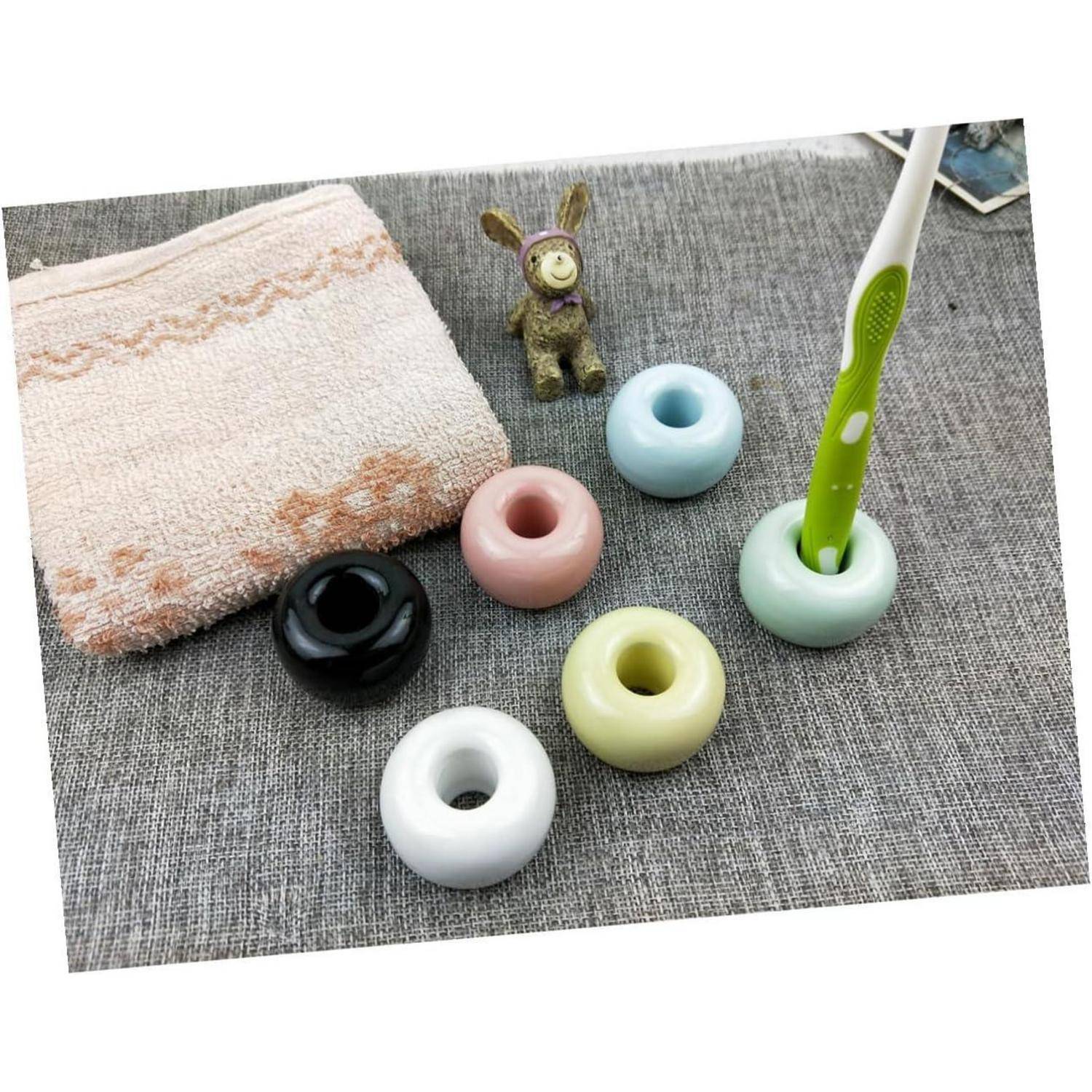 Eine Zahnbürste steht in einem mintgrünen Ring neben mehreren pastellfarbenen, ringförmigen Haltern, einem kleinen Spielzeugkaninchen und einem gefalteten Handtuch auf einer grauen Matte.