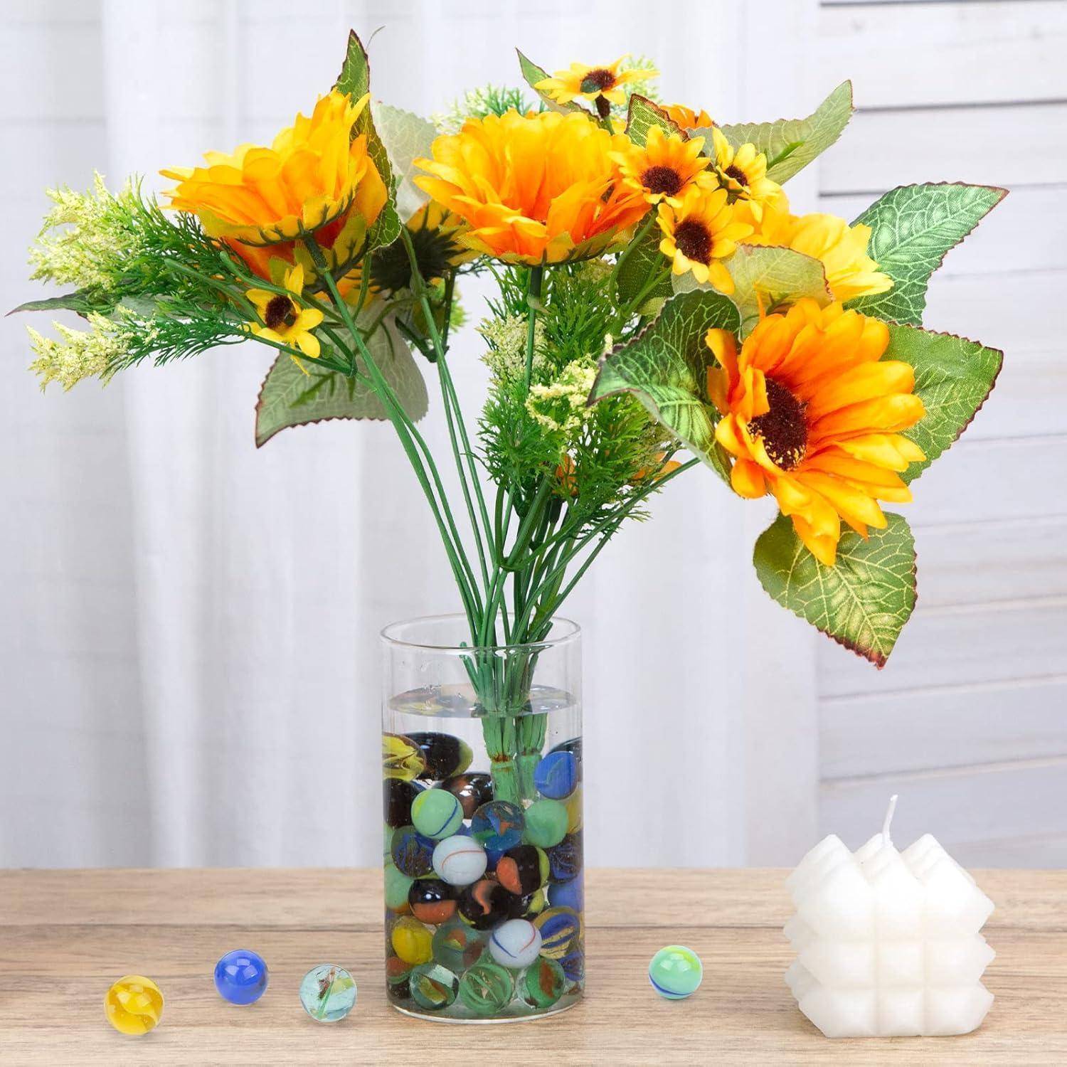 Sonnenblumenstrauß in einer Glasvase gefüllt mit bunten Murmeln auf einem Holztisch, neben einer weißen Kerze.