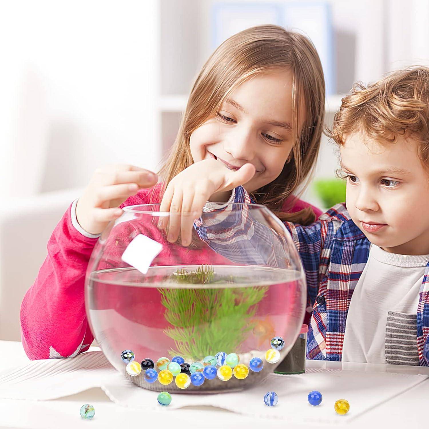 Zwei Kinder schauen sich ein Aquarium mit einem kleinen Fisch und bunten Murmeln auf einem Tisch an.
