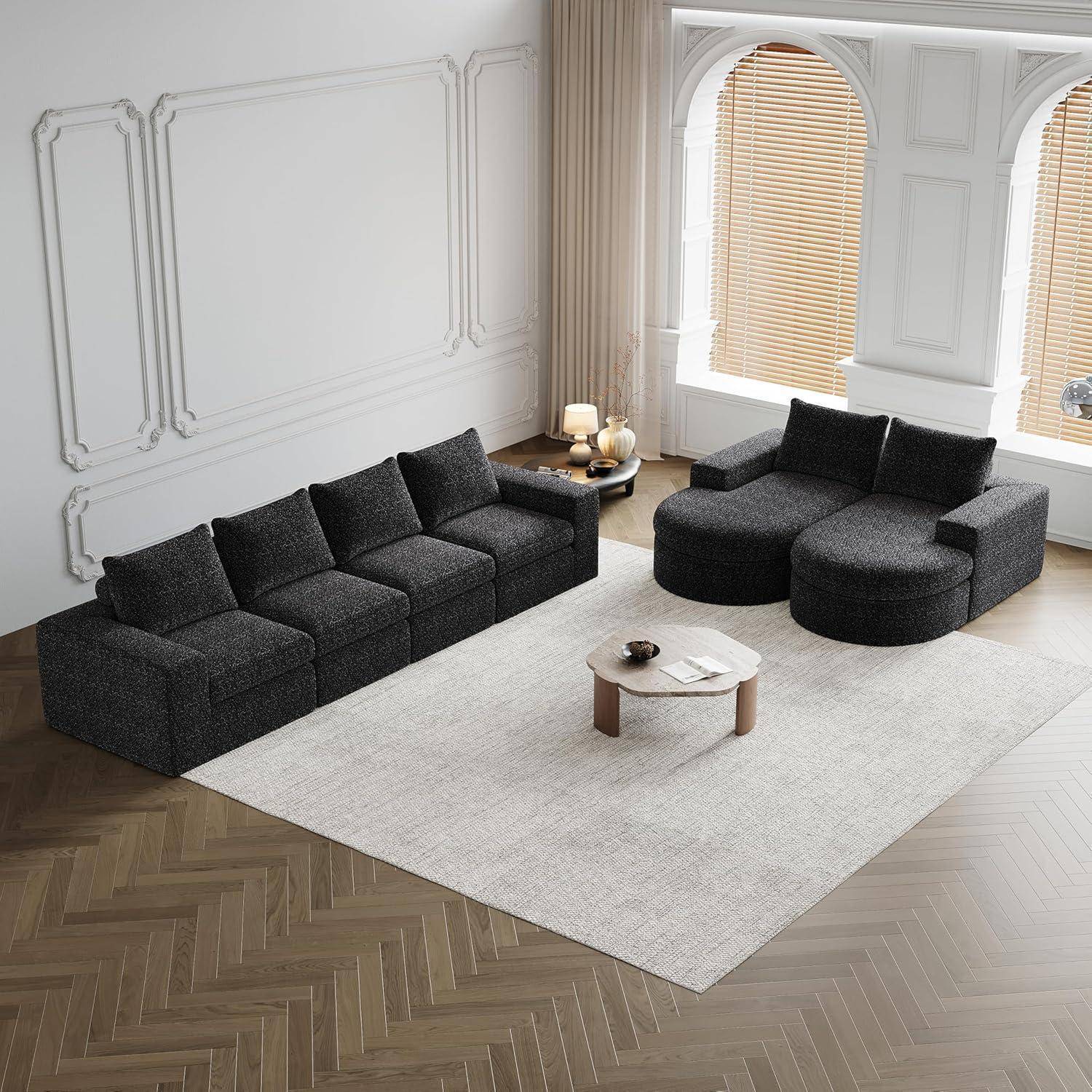 Modernes Wohnzimmer mit dunklem modularem Ecksofa und runden Chaise-Sofas auf einem hellen Teppich, zentriert um einen niedrigen Couchtisch in der Nähe großer gewölbter Fenster mit Jalousien.