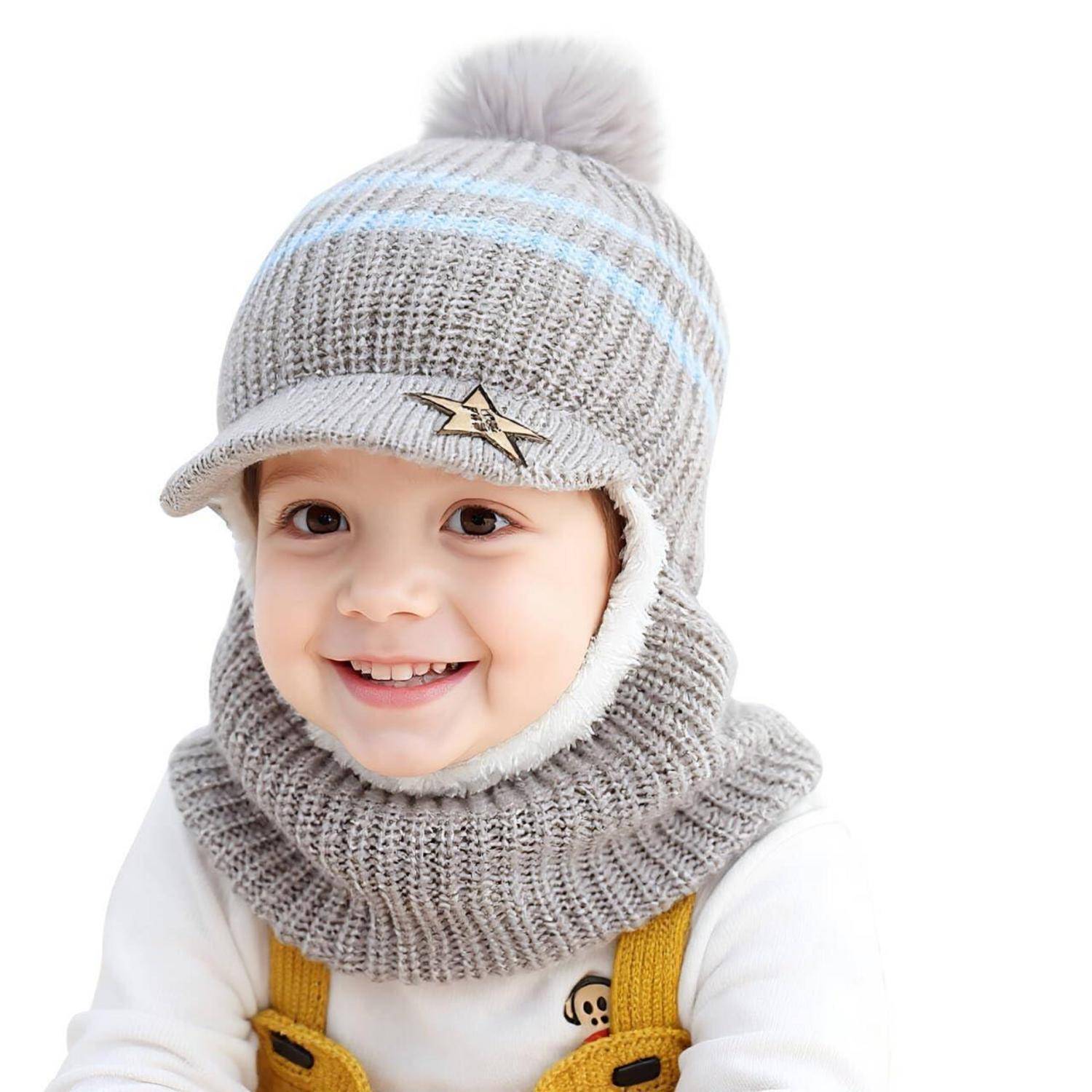 Lachendes Kleinkind, das eine graue Strickmütze mit Pompon und einen passenden Schal über einem weißen Hemd und gelben Hosenträgern auf weißem Hintergrund trägt.