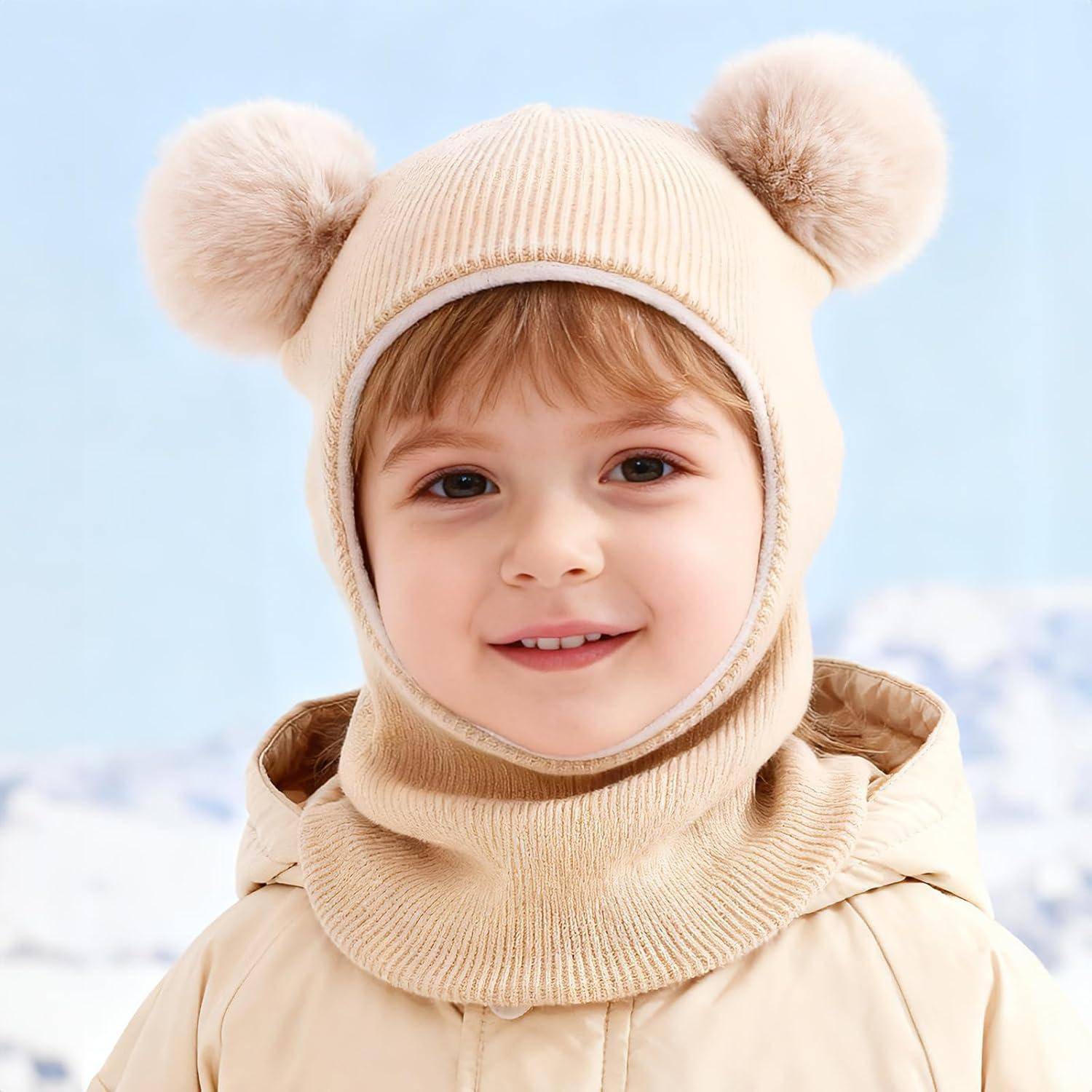 Lachendes junges Kind in einer beigen Wintermütze mit Bommel-Ohren und passendem Mantel vor einem verschneiten, hellblauen Hintergrund.
