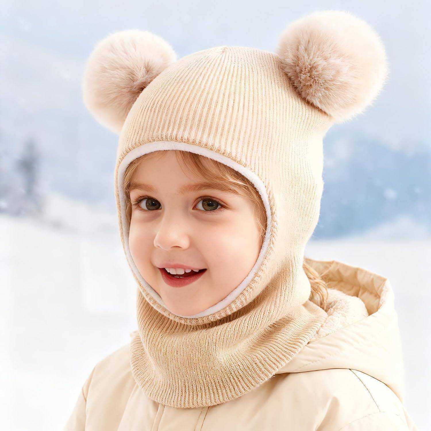 Lächelndes Kind in einer beigen Wintermütze mit Pompon-Ohren und passendem Kapuzenschal, posiert in einer verschneiten Landschaft im Freien.