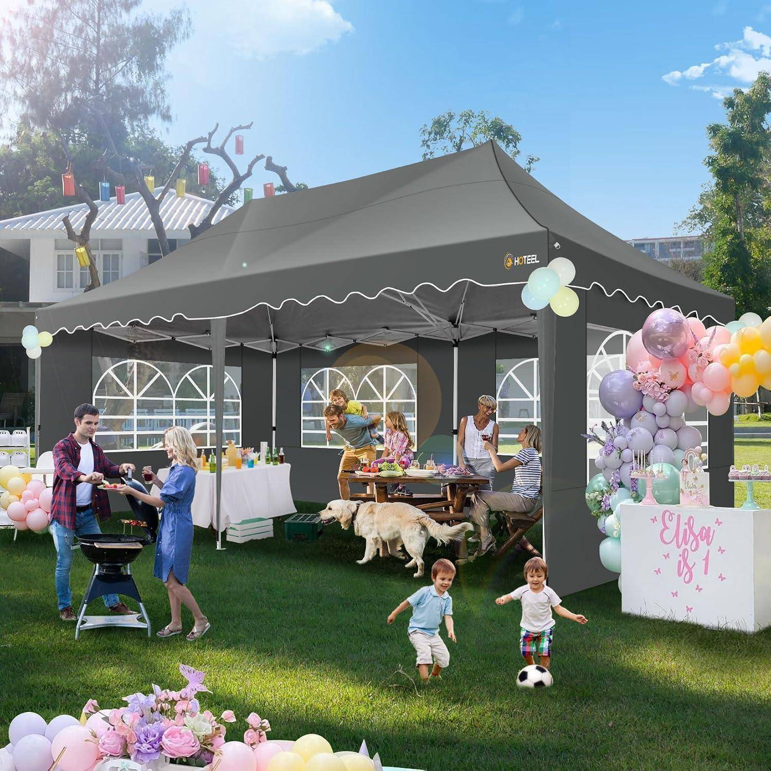 Geburtstagsfeier im Freien unter einem schwarzen Pavillon-Zelt, mit Erwachsenen, die Essen servieren, Kindern, die auf dem Gras spielen, und Pastellballon-Dekorationen neben einem Schild mit der Aufschrift „Elsa ist 1