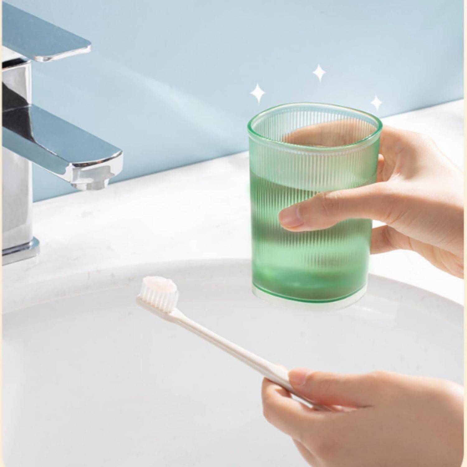 Hände halten ein grünes Glas Wasser und eine Zahnbürste an einem Waschbecken im Badezimmer neben einem Wasserhahn.