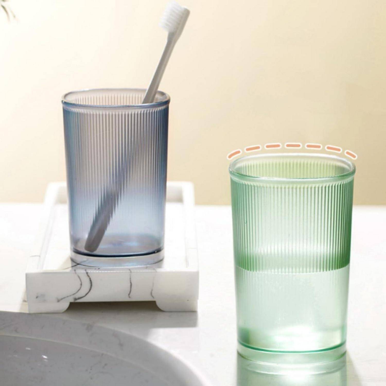 Zwei gerippte, durchscheinende Becher auf einer Spülbeckenablage: ein blauer Becher mit einer Zahnbürste und ein grüner Becher mit orange hervorgehobener Krempe.