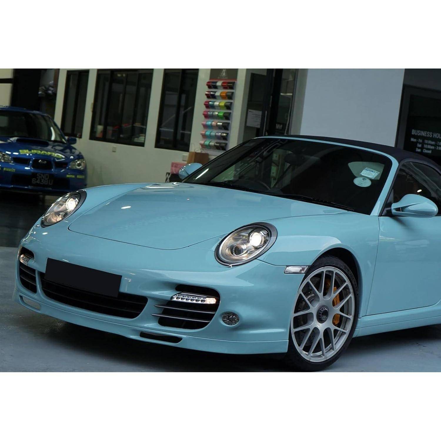 Hellblauer Porsche-Sportwagen in Innenräumen geparkt, Vorderansicht von links mit schwarzem Verdeck und silbernen Rädern.