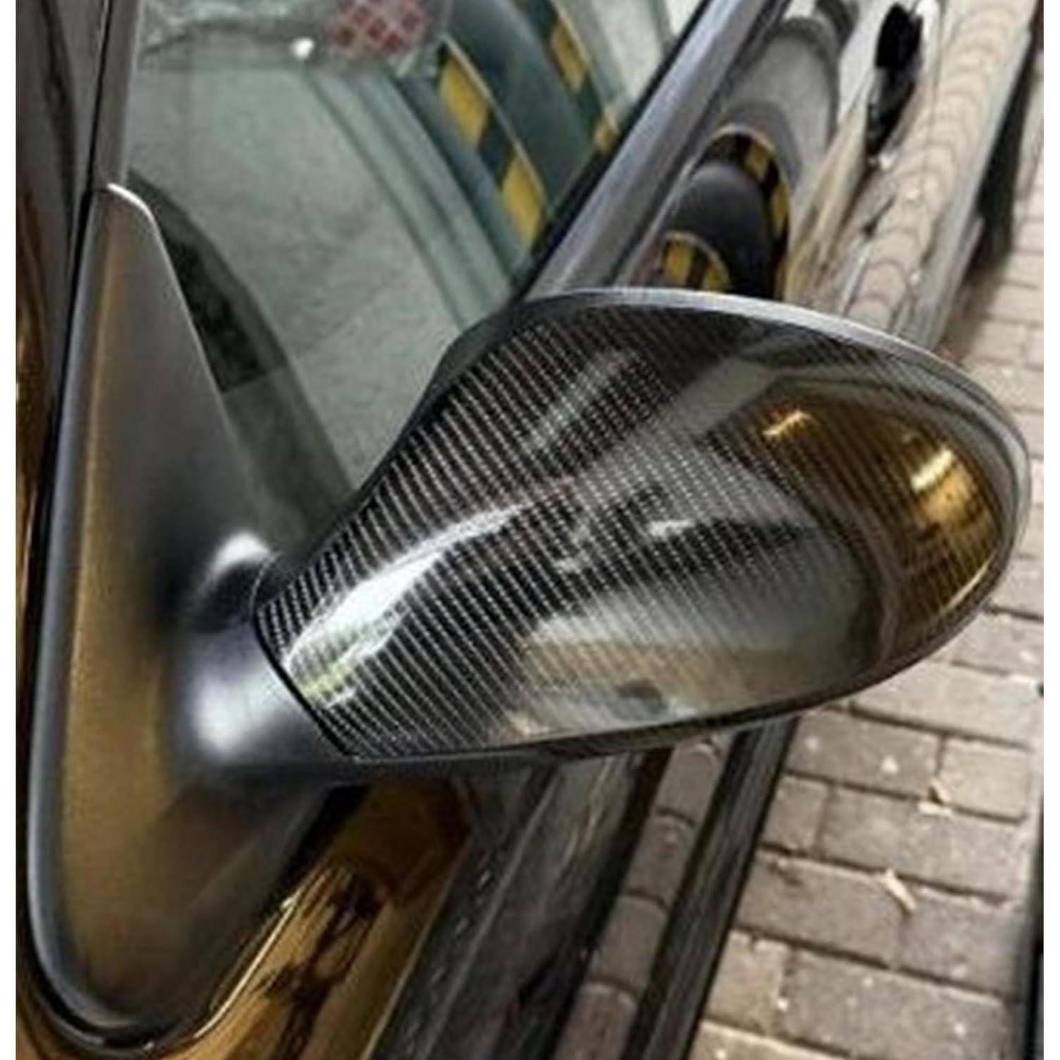 Nahaufnahme eines Seitenspiegels eines Autos mit Kohlefaser-Muster, angebracht an einem dunklen Fahrzeug, das neben einem Gehweg aus Ziegeln geparkt ist.
