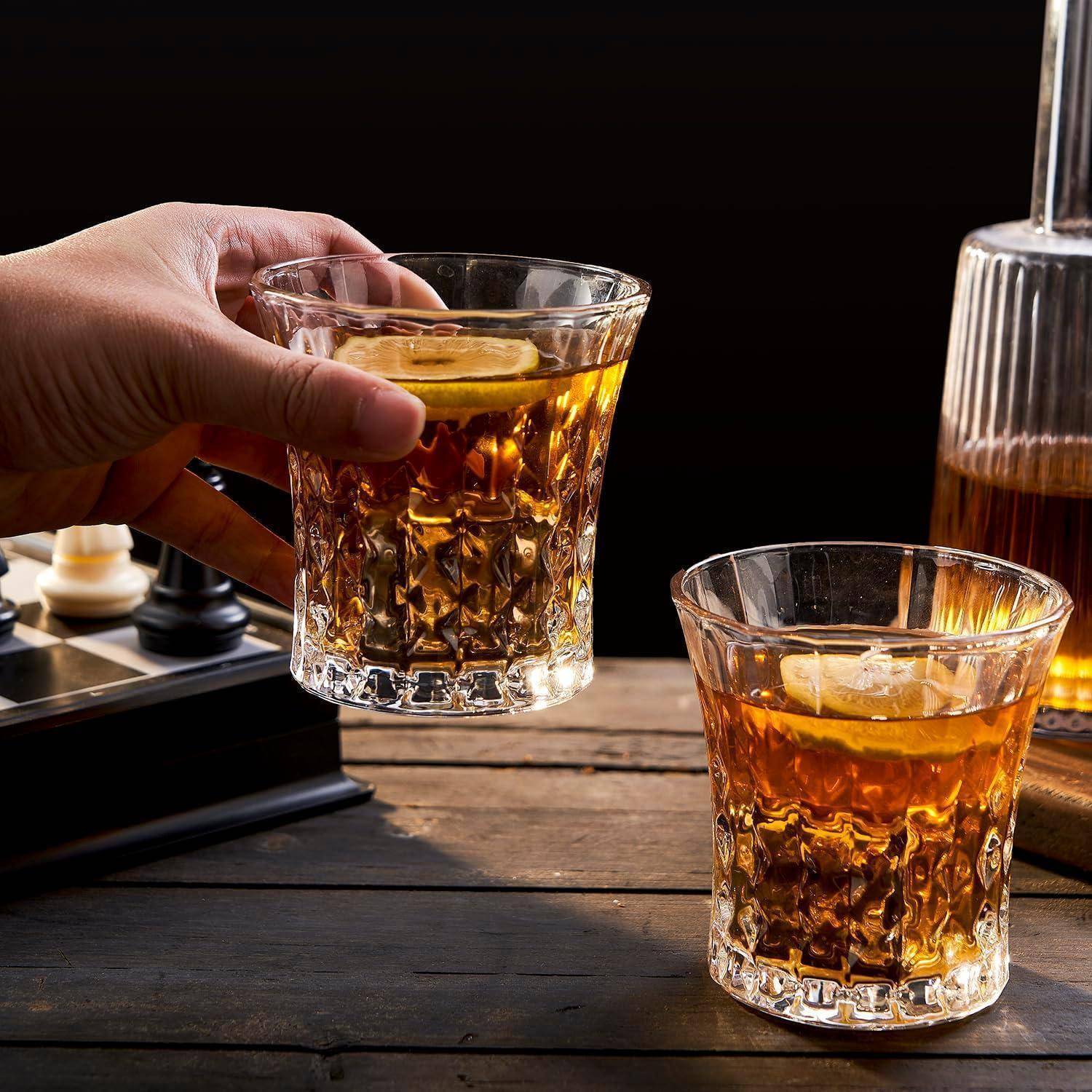 Eine Hand hält ein Glas mit bernsteinfarbenem Whiskey mit Eis und einer Zitronenscheibe neben einem ähnlichen Getränk auf einem Holztisch.