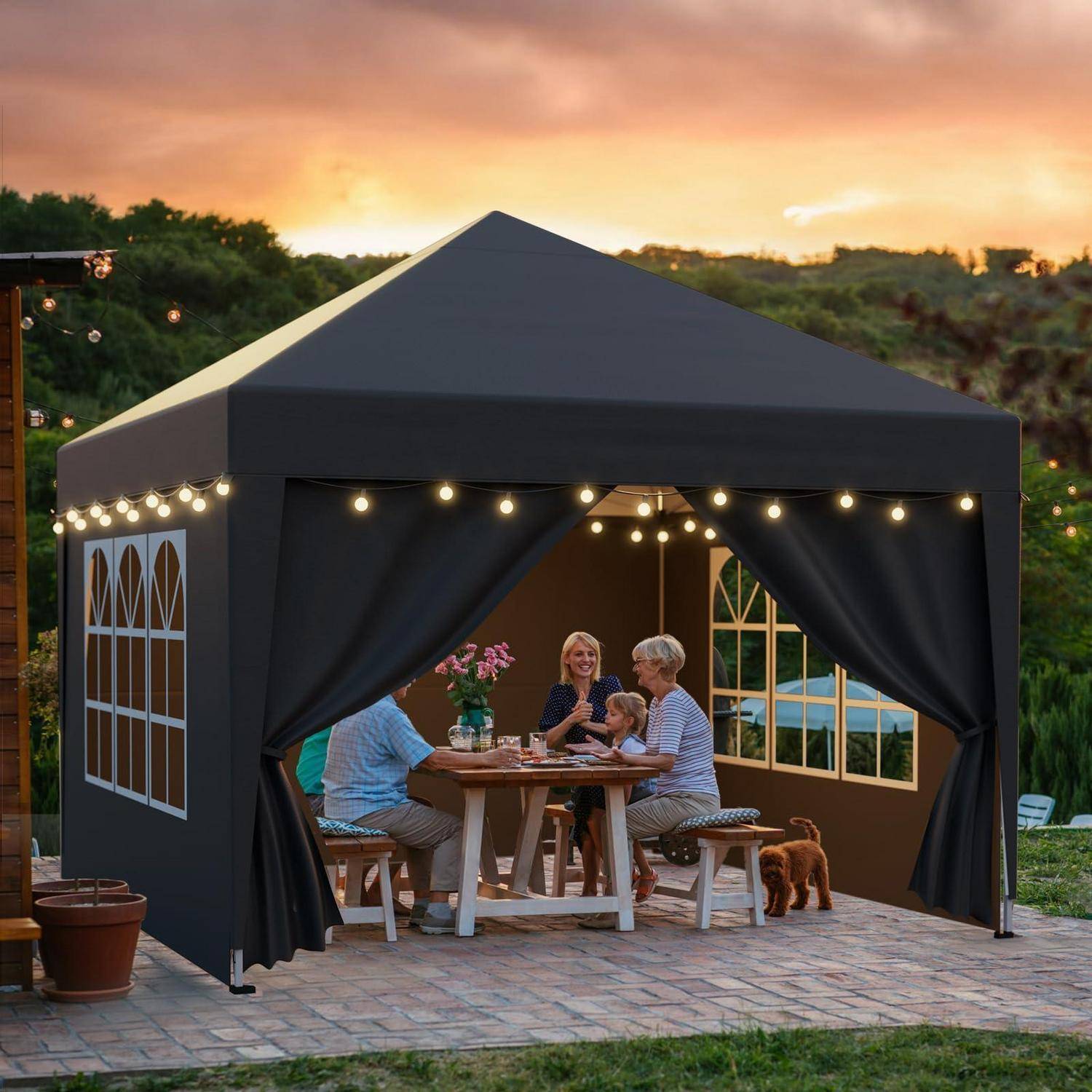 Familie sitzt an einem Picknicktisch in einem schwarzen Außenpavillonzelt bei Sonnenuntergang, mit Lichterketten und einem kleinen Hund auf einer Terrasse.