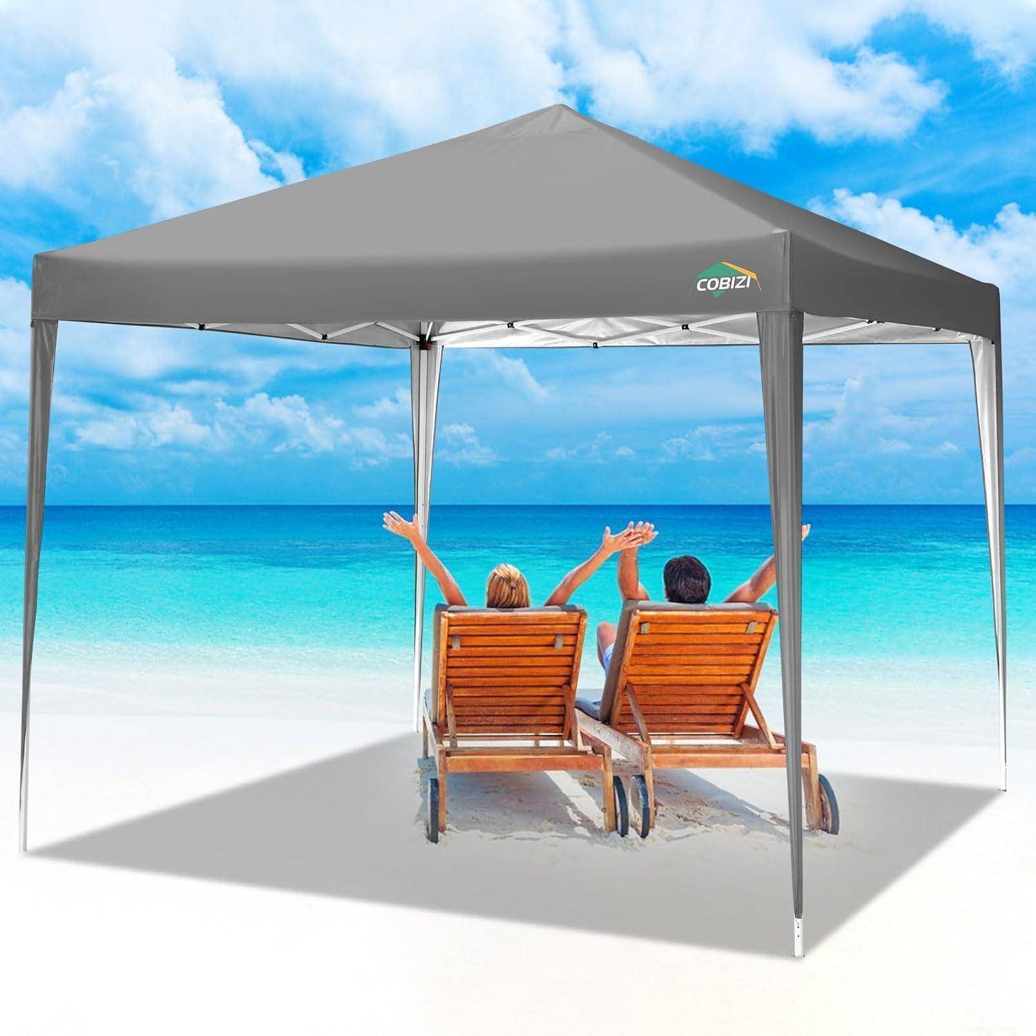 Zwei Personen entspannen sich in Holzstrandkörben unter einem grauen Pop-up-Dach und blicken auf den Ozean mit erhobenen Armen an einem sonnigen Strand.