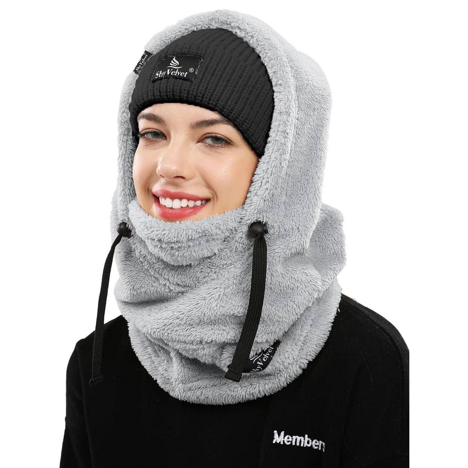 Lächelnde Frau, die eine graue Fleece-Kapuze und einen Nackenerwärmer über einer schwarzen Beanie und einem schwarzen Oberteil trägt, vor einem weißen Hintergrund gezeigt.