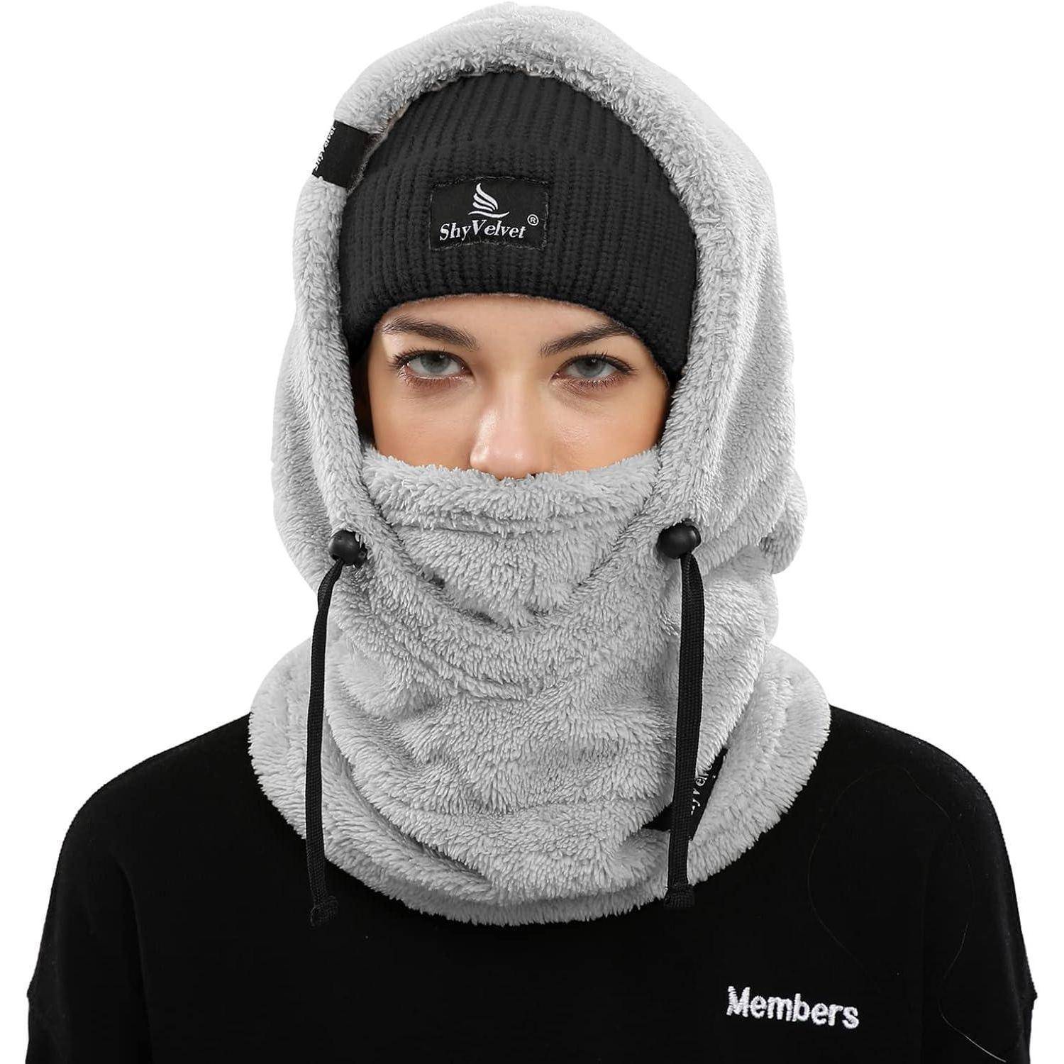 Person, die einen grauen Fleece-Kapuzenpulli und eine Gesichtsmaske über einer schwarzen Mütze trägt, wobei nur die Augen vor einem einfachen weißen Hintergrund sichtbar sind.