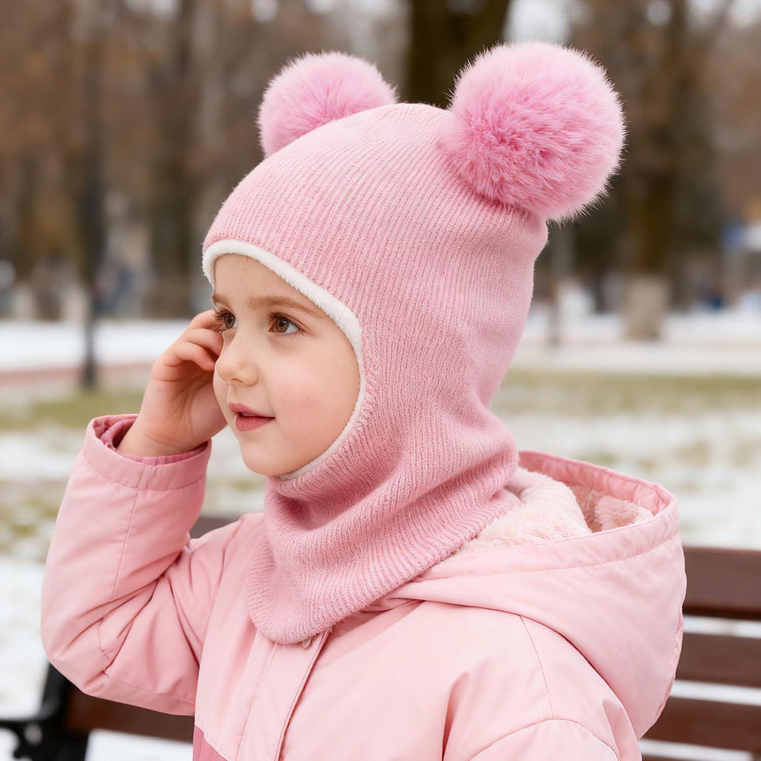 Kleines Kind in einer rosa Wintermütze und einem Mantel sitzt auf einer Bank im Freien und fasst sich ans Ohr in einem verschneiten Park.