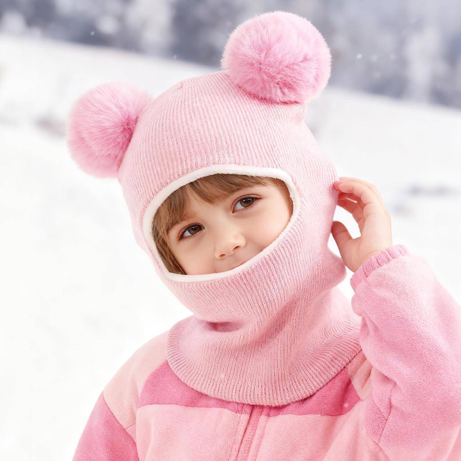 Kind in einer rosa Wintermütze mit Bommel, das in die Kamera schaut und die Kapuze in einer verschneiten Außenumgebung zurechtzieht.