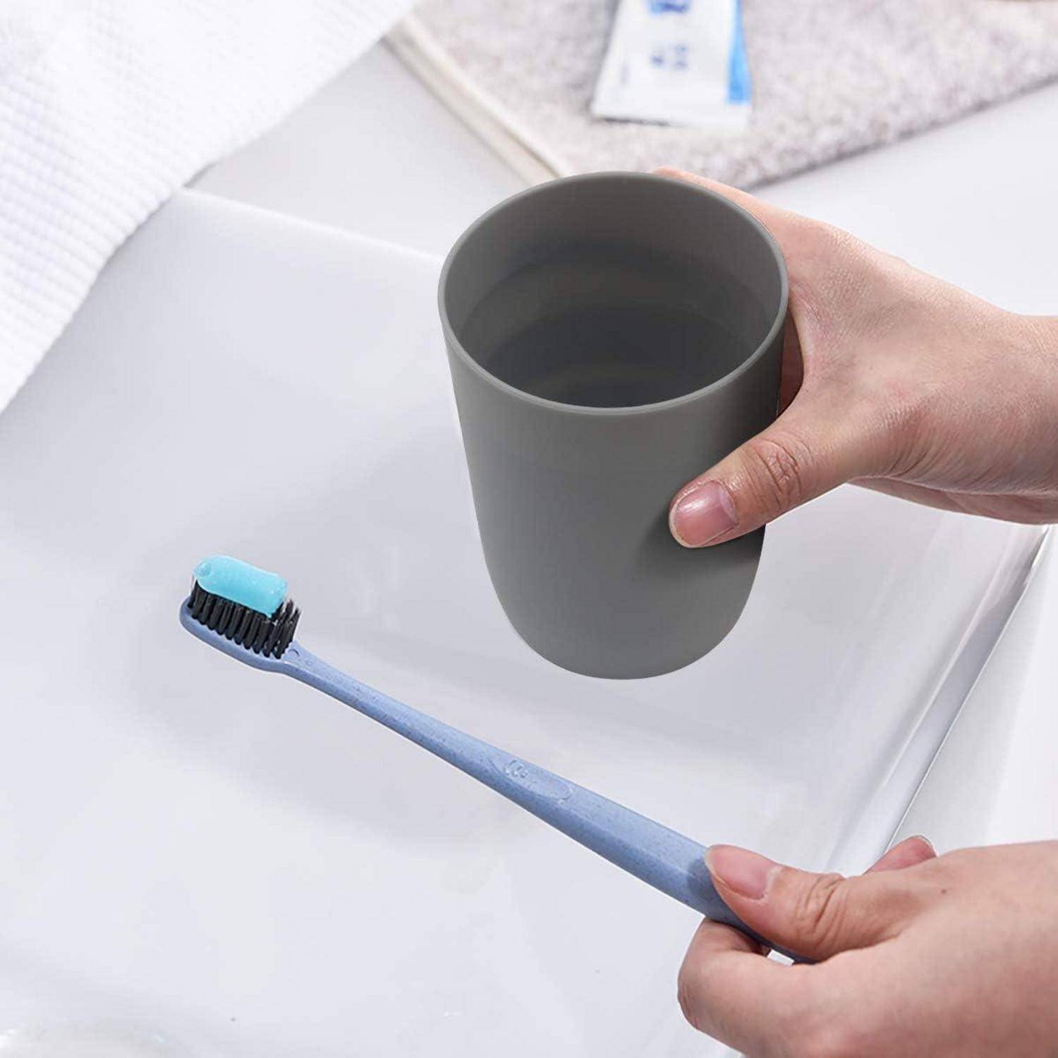 Hände halten eine graue Tasse über einer blauen Zahnbürste mit Paste auf einer weißen Oberfläche, was auf einen Badezimmer-Hygiene-Kontext hindeutet.
