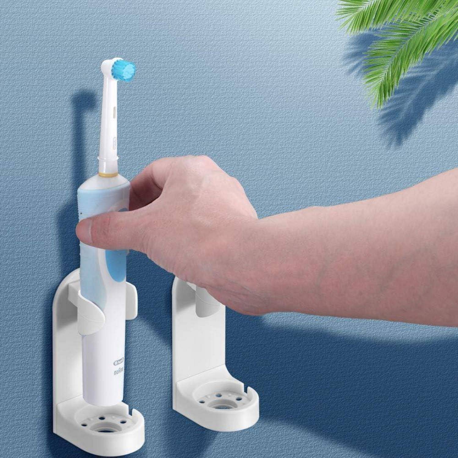 Hand, die eine elektrische Zahnbürste in eine an der Wand montierte Ladeschale neben einem zweiten leeren Halter gegen eine blaue Wand einlegt.