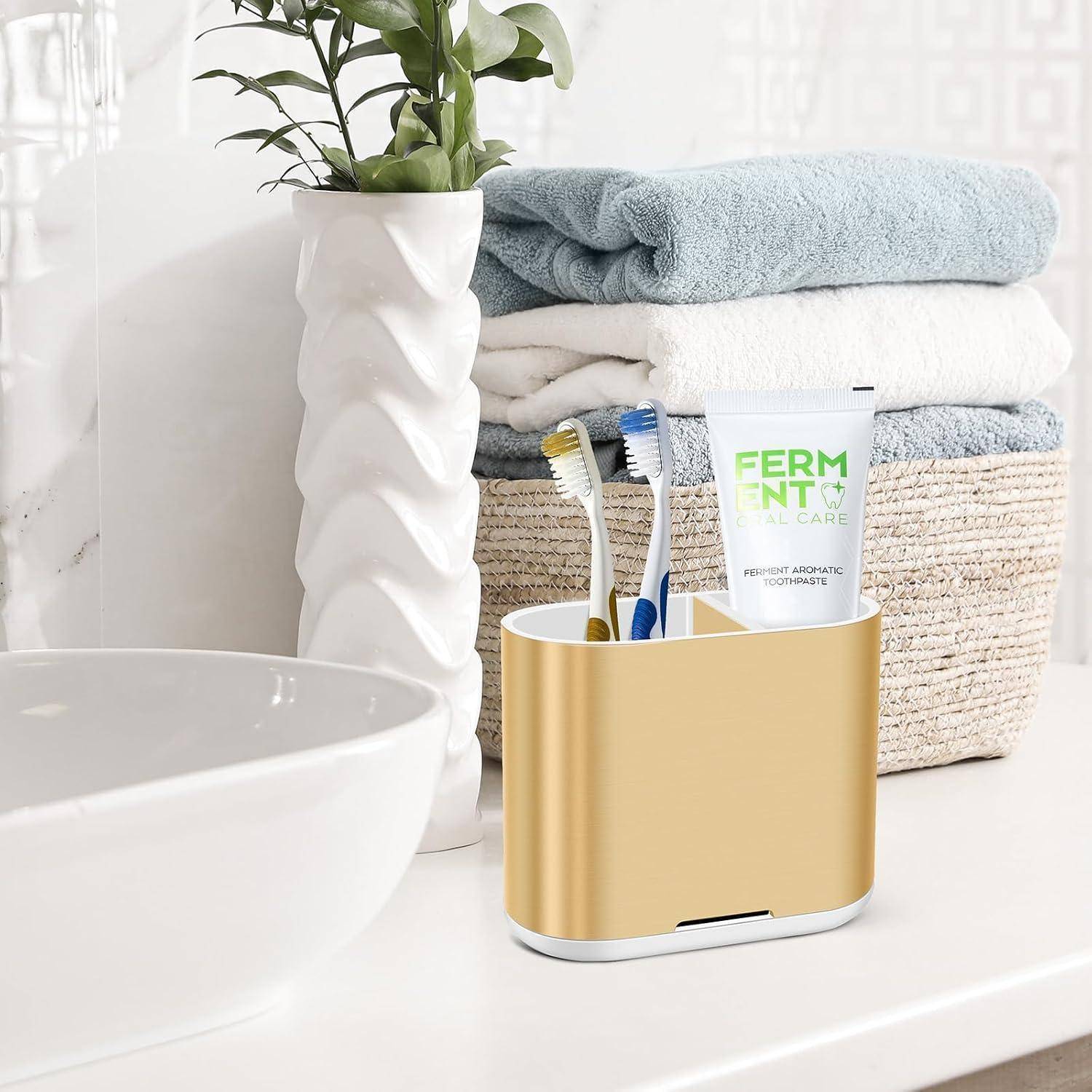 Goldener Badezimmer-Arbeitsplatte-Organizer mit Zahnbürsten und Zahnpasta neben gestapelten Handtüchern und einer Pflanze neben einem Waschbecken.