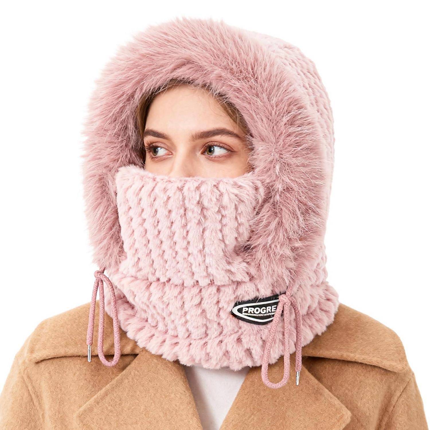 Person trägt eine rosa pelzgefütterte Kapuze und Gesichtsbedeckung, wobei nur die Augen sichtbar sind, und einen beigen Mantel vor weißem Hintergrund.