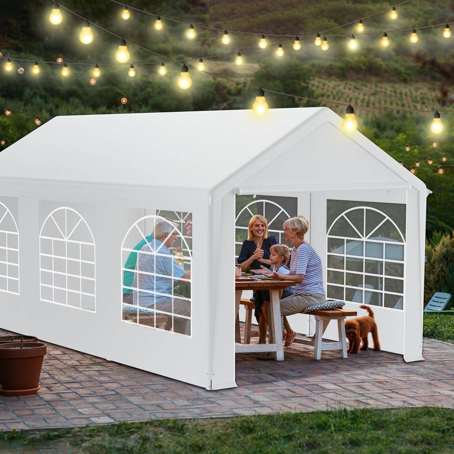 Familie isst in einem weißen Außenzelt auf einer Ziegelpflaster-Terrasse, mit Lichterketten über Kopf und einem kleinen Hund neben dem Tisch.