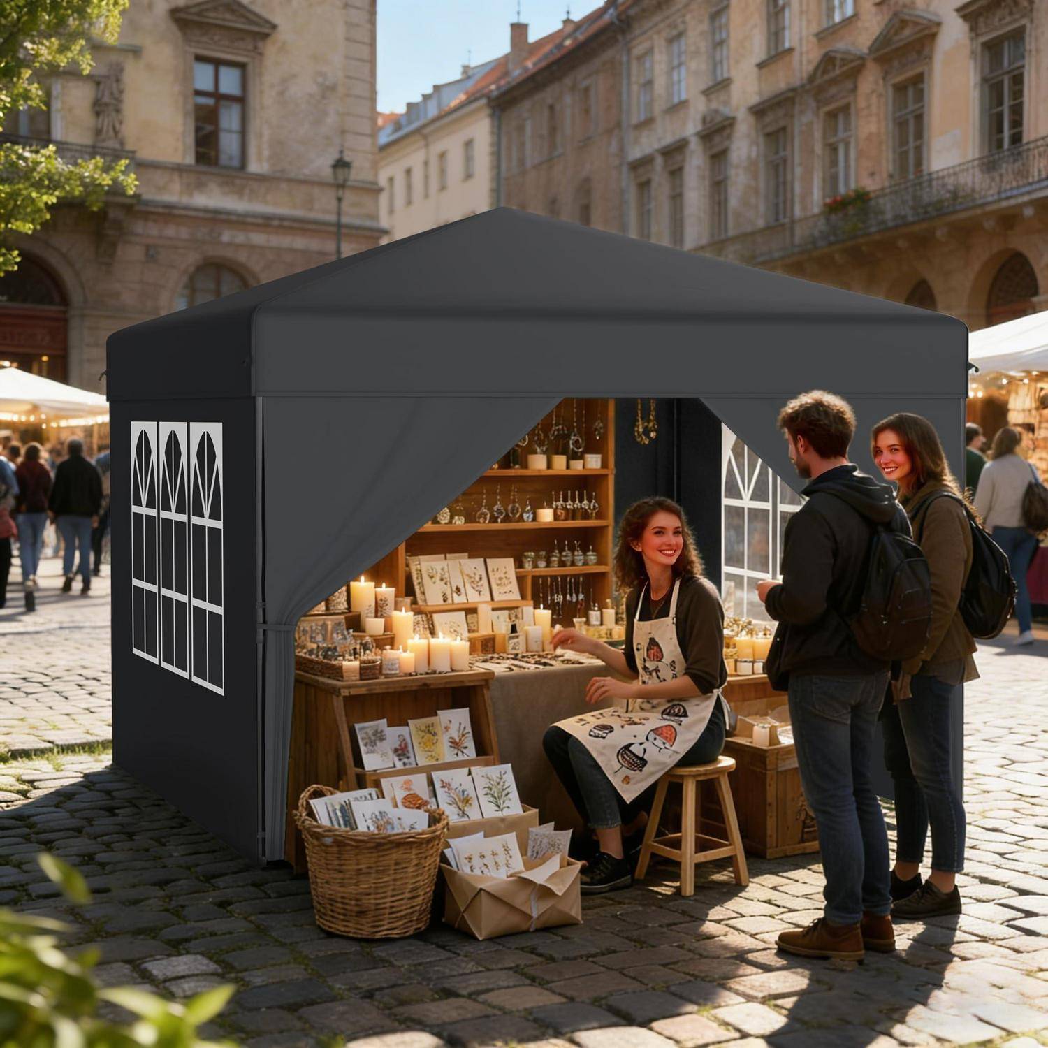 Marktstand im Freien unter einem schwarzen Pavillon-Zelt, mit einem Verkäufer, der handgefertigte Karten und Kerzen an zwei Kunden auf einem Kopfsteinpflaster-Platz verkauft.