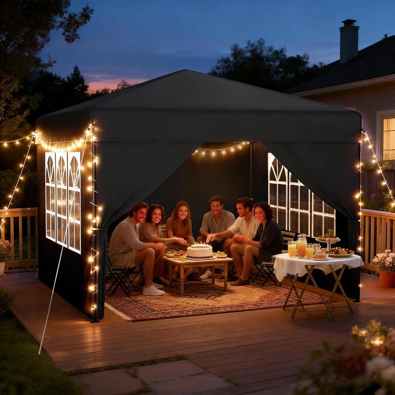 Freunde versammelten sich in einem beleuchteten Baldachinzelt im Hinterhof in der Abenddämmerung und feierten um einen Kuchen auf einer Terrasse mit Getränken und einem Beistelltisch.