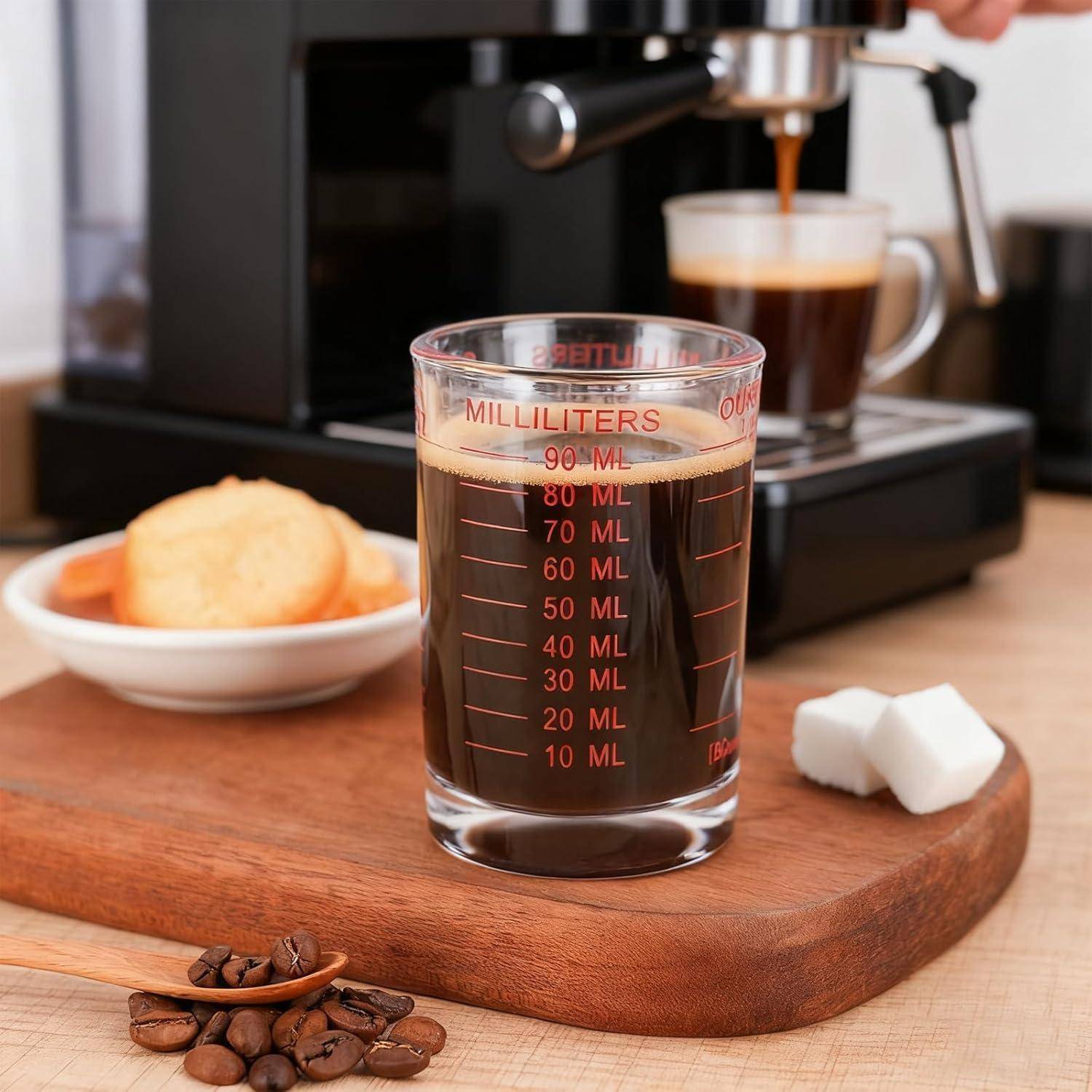 Messbehälter mit Milliliterskala gefüllt mit Kaffee neben einer Espressomaschine, mit einem Gebäck, Zuckerwürfeln und Kaffeebohnen auf einem Holztablett.