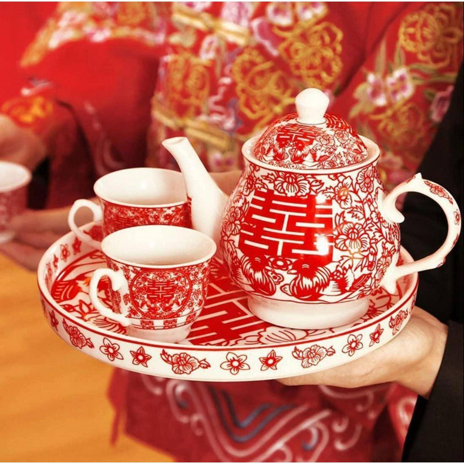 Hand hält ein rot-weiß bemaltes Porzellan-Teeservice mit Teekanne, Tassen und Tablett, das mit Doppelglück- und floralen Hochzeitsmotiven verziert ist.