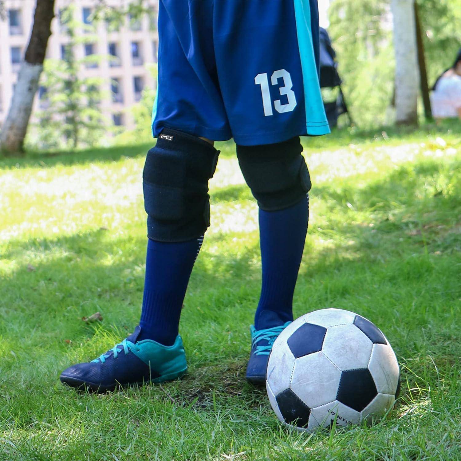 Kind in blauer Fußballuniforms auf Gras neben einem Fußball stehend, mit sichtbaren Knieschonern und Trikotnummer 13.