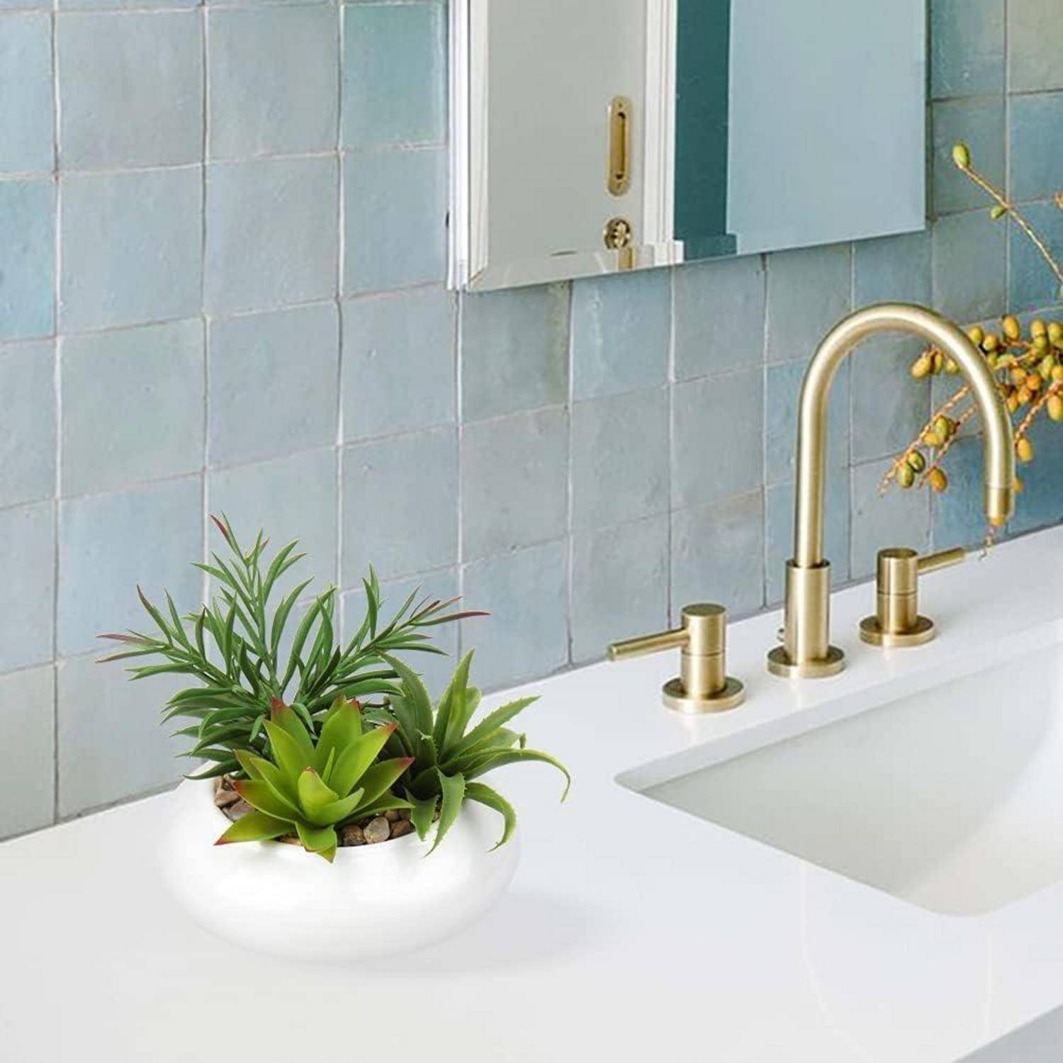 Weiße Badezimmerkommode mit goldenem Wasserhahn, Waschbecken und kleinen Töpfen mit Sukkulenten vor einer blaugeflieselten Wand.