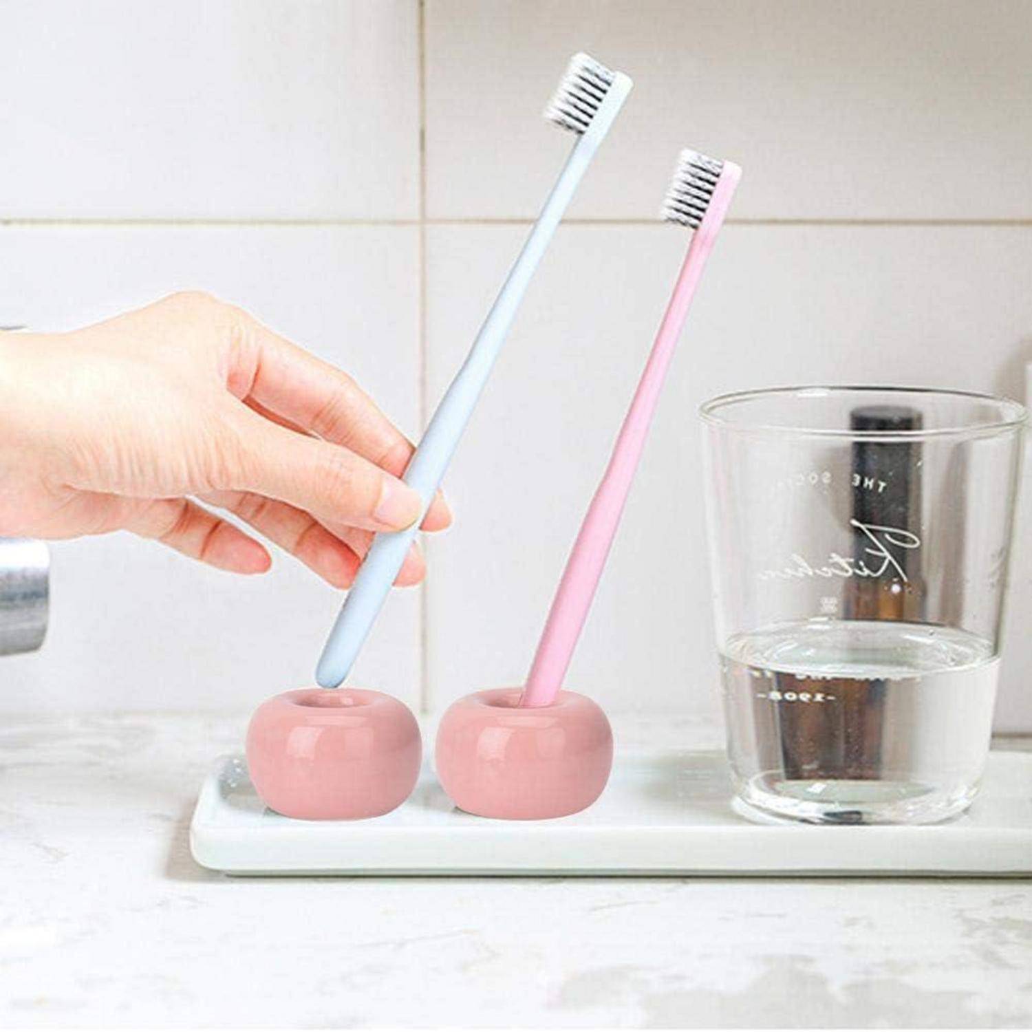 Eine Hand platziert eine hellblaue Zahnbürste in einem rosa Halter neben einer rosa Zahnbürste und einem Glas Wasser auf einer Badezimmerablagefläche.