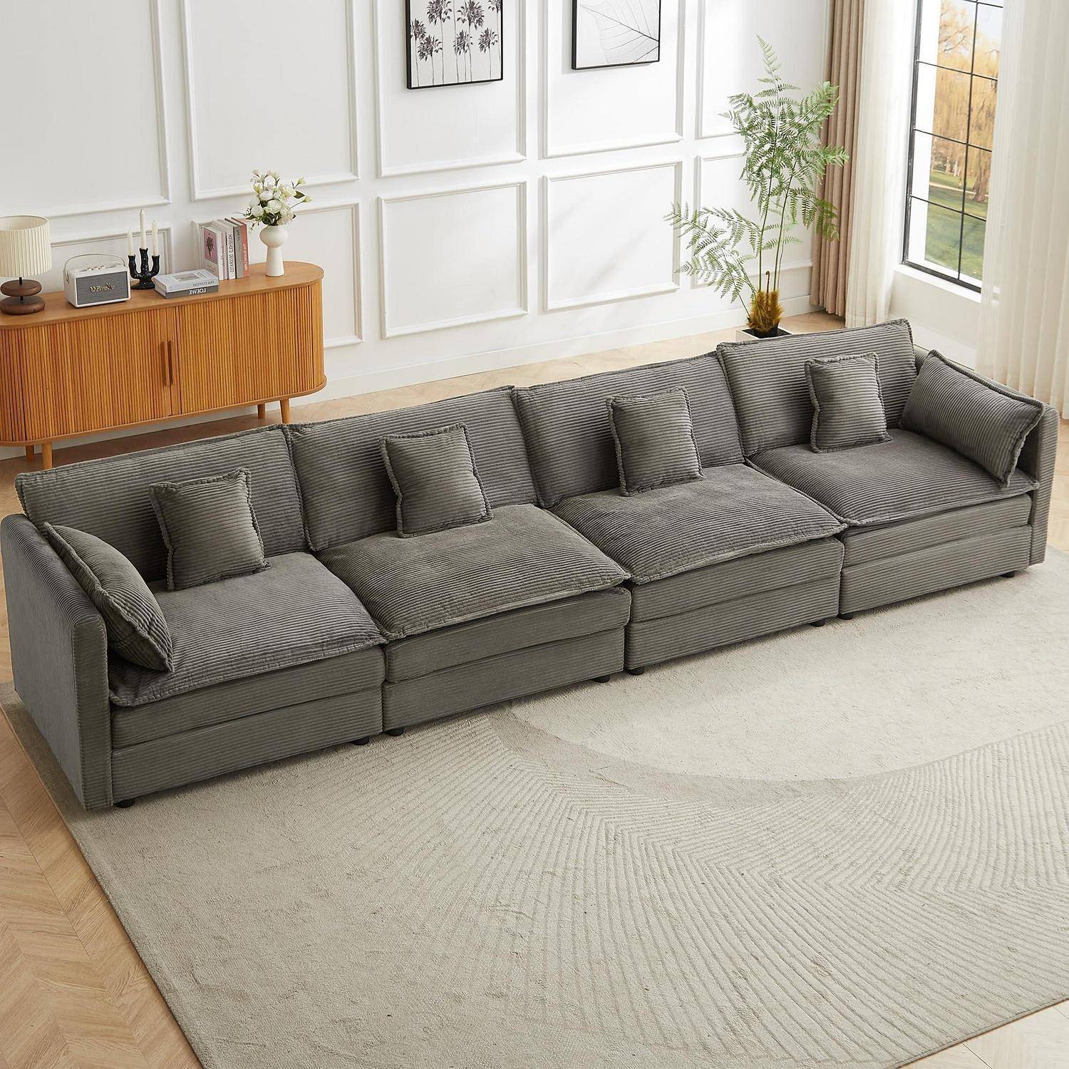 Graues modulares Ecksofa mit fünf Rückenkissen und quadratischen Dekokissen in einem hellen Wohnzimmer neben einem Holzbeistell-/Sideboard, einem Teppich und einem Fenster.