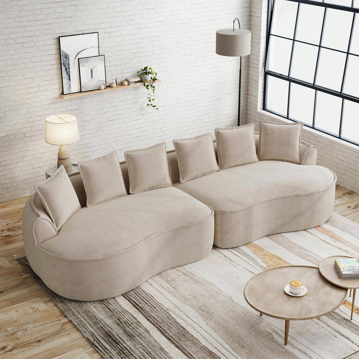 Beiges modulares geschwungenes Sofa mit fünf Kissen in einem hellen Wohnzimmer, kombiniert mit einem runden Couchtisch, einer Stehleuchte, einem Wandregal und einem großen Fenster.