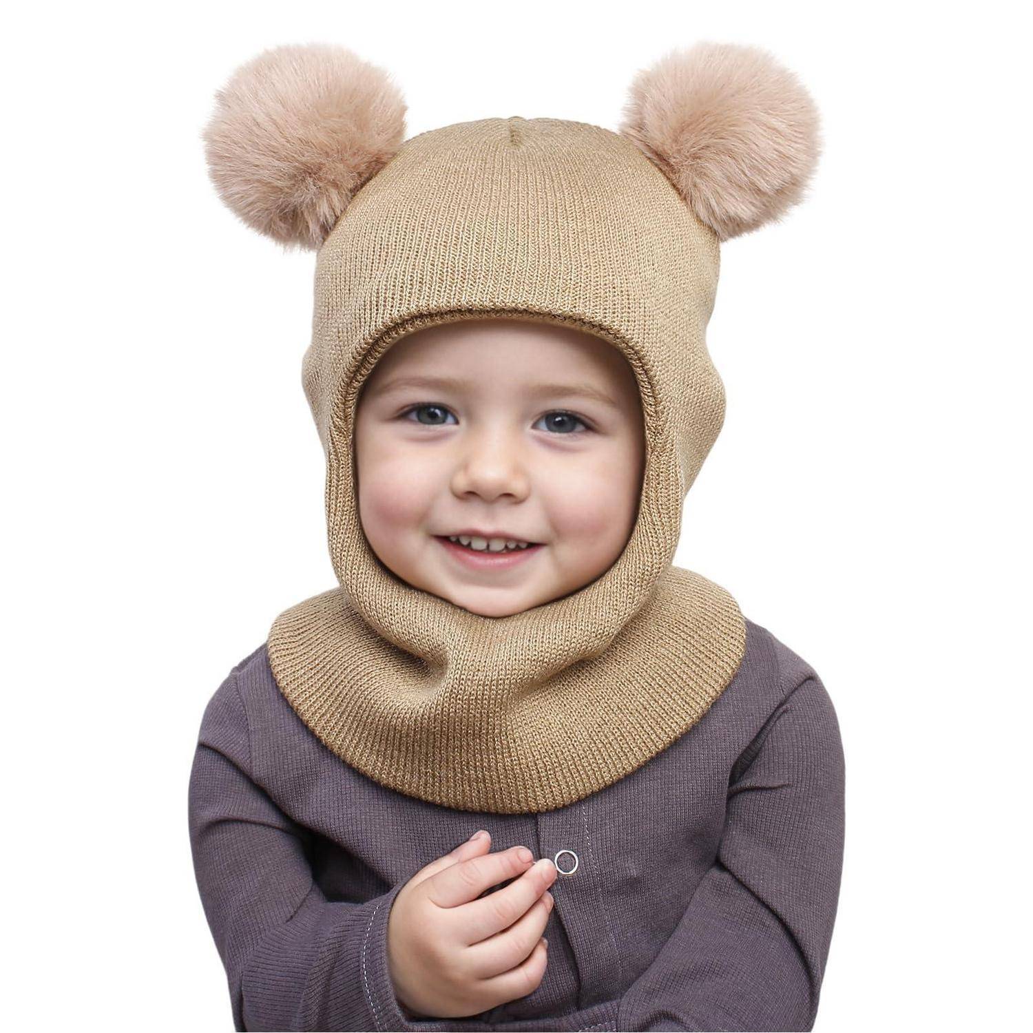 Lächelndes Kleinkind mit einer beigefarbenen Strickmütze mit Bärenohren und passendem Schal, das einen kleinen Ring gegen einen weißen Hintergrund hält.