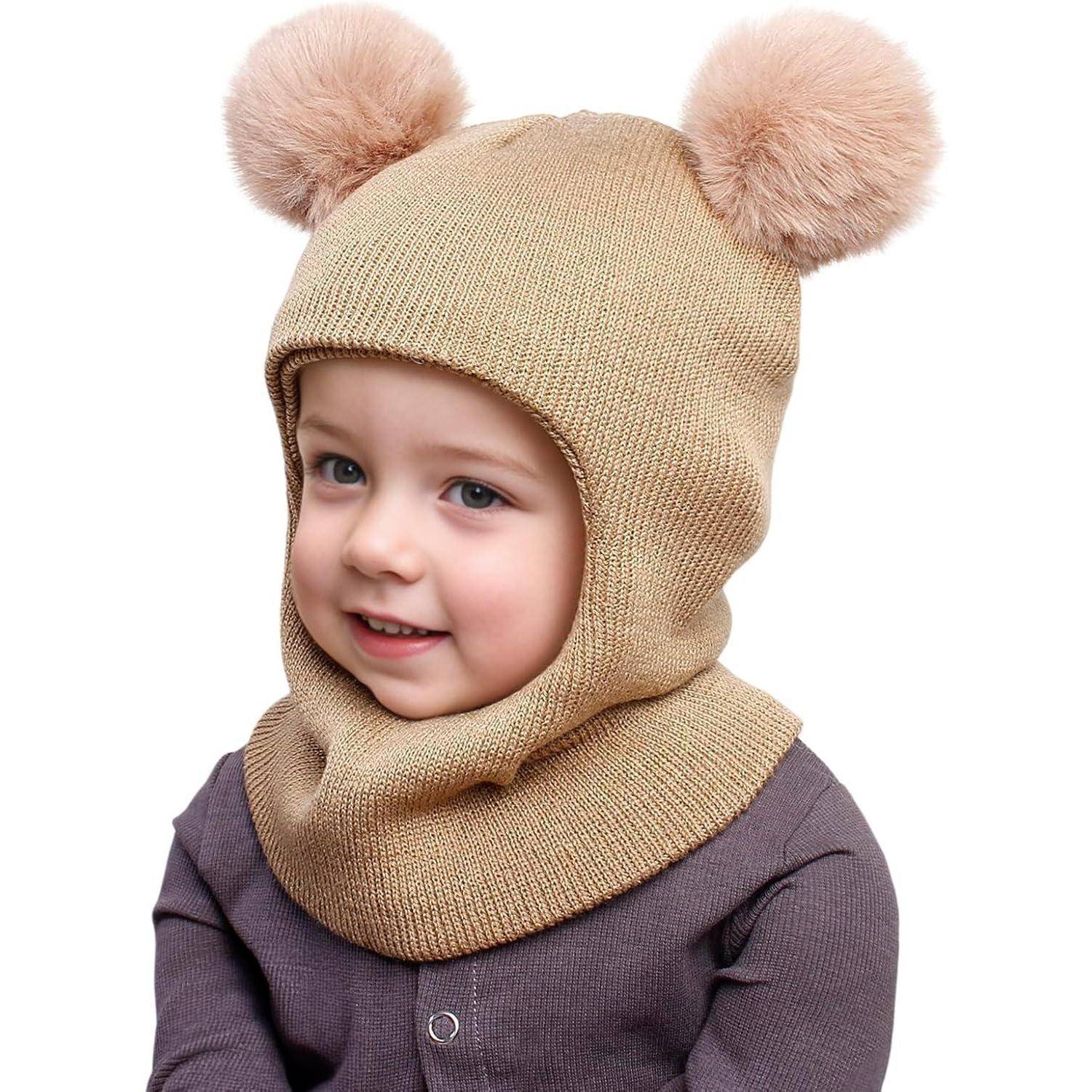 Kleines Kind, das eine beige Strickbalacklava mit zwei Pompon-Ohren trägt, lächelnd vor weißem Hintergrund.