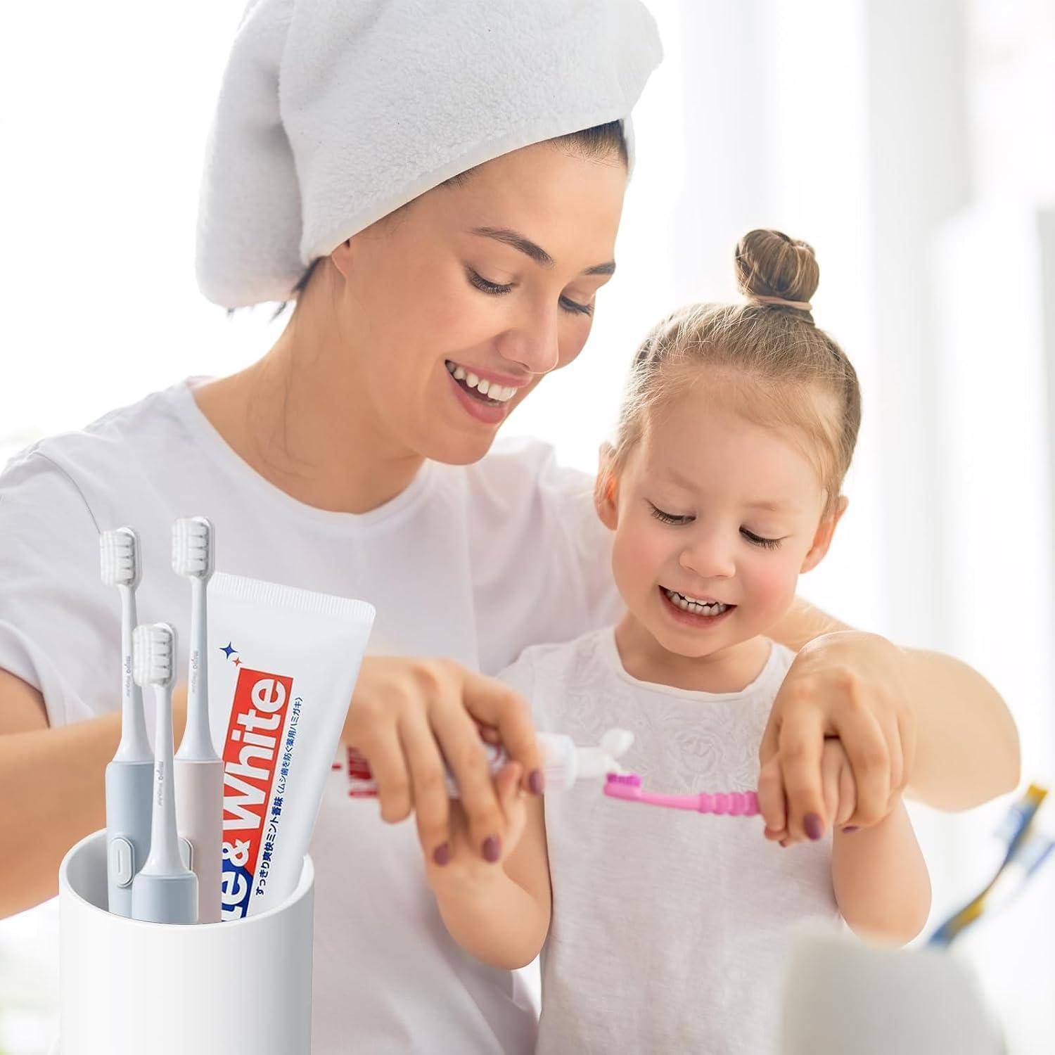 Lächelnde Mutter hilft ihrer jungen Tochter beim Zähneputzen, mit elektrischen Zahnbürsten und sichtbarer Colgate-Zahnpasta in einem hellen Badezimmer.