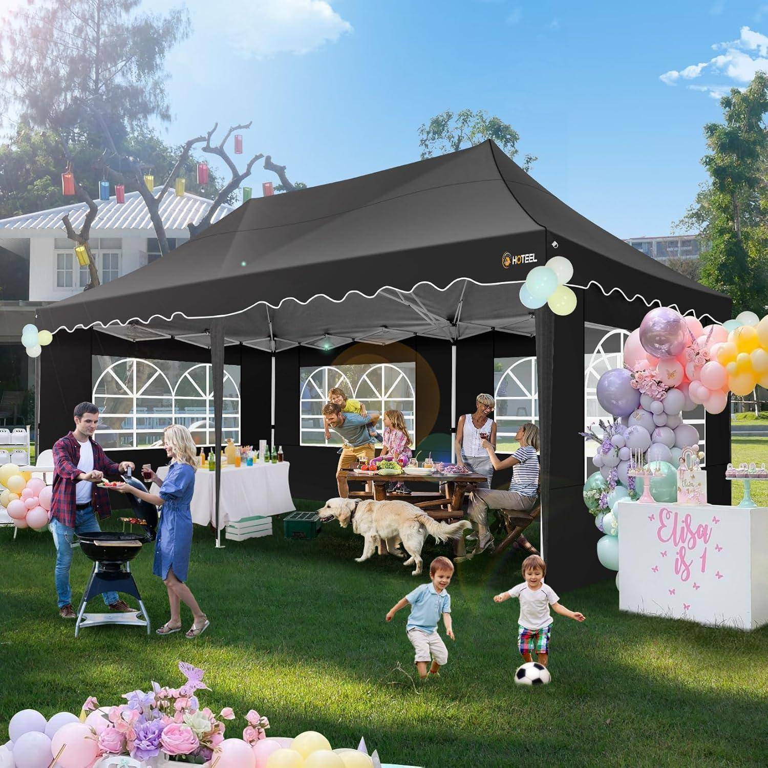 Outdoor-Geburtstagsfeier unter einem schwarzen Partyzelt, mit Erwachsenen, die Essen servieren, Kindern, die auf dem Rasen spielen, Hunden in der Nähe und Dekoration mit Pastellballons.