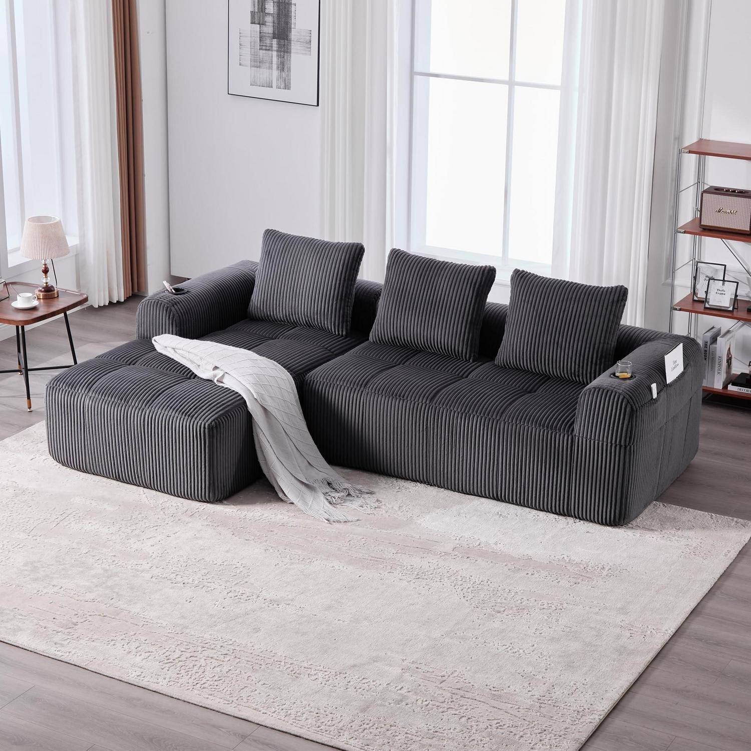 Modernes Wohnzimmer mit einem dunkelgestreiften Ecksofa, drei Kissen und einer hellen Wurfdecke auf einem großen weißen Teppich neben hellen Fenstern.