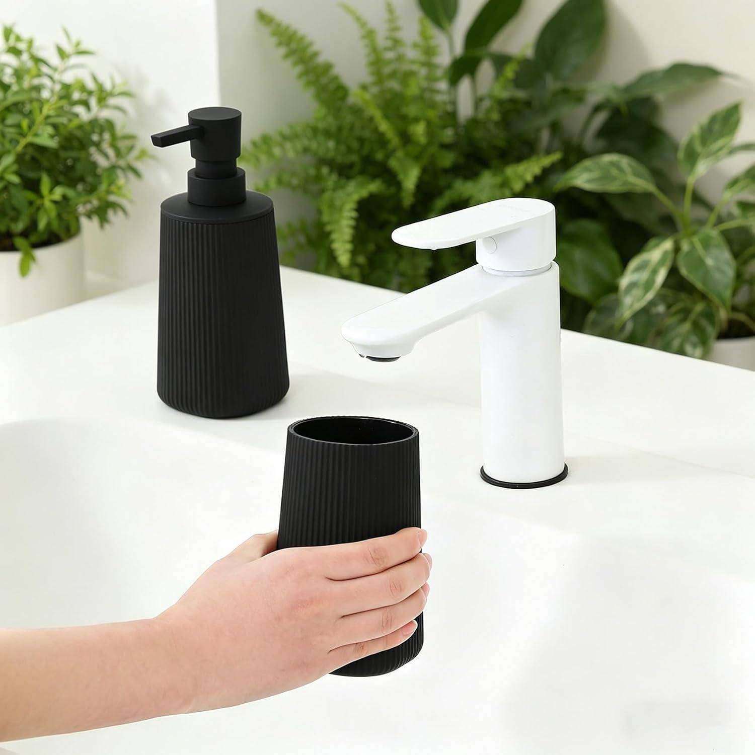 Eine Hand stellt einen schwarzen gerippten Becher auf eine weiße Arbeitsplatte neben einen schwarzen Seifenspender und einen weißen Wasserhahn, mit grünen Pflanzen im Hintergrund.