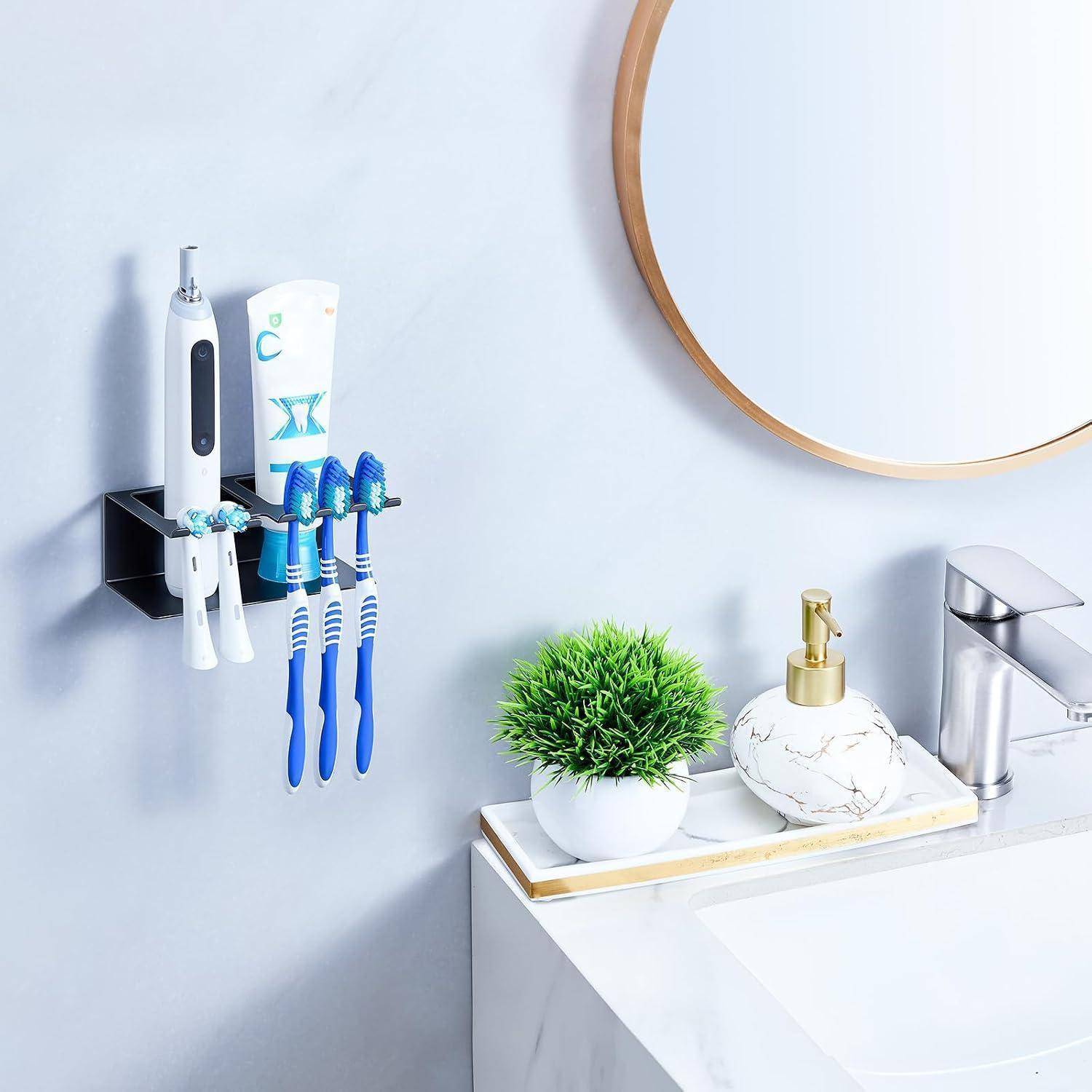 Badezimmerwaschbeckenbereich mit einem wandmontierten Halter für eine elektrische Zahnbürste, Zahnpasta und drei manuelle Zahnbürsten neben einem Spiegel, einer Pflanze und einem Seifenspender.