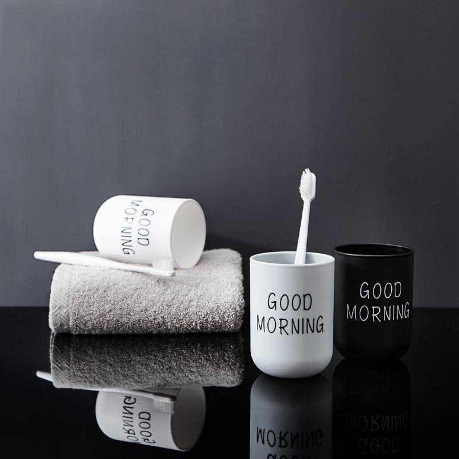 Minimalistische Badezimmer-Stillleben mit zwei 'GUTEN MORGEN'-Zahnburstbechern, einer Zahnbürste und gefalteten Handtüchern auf einer reflektierenden Oberfläche.