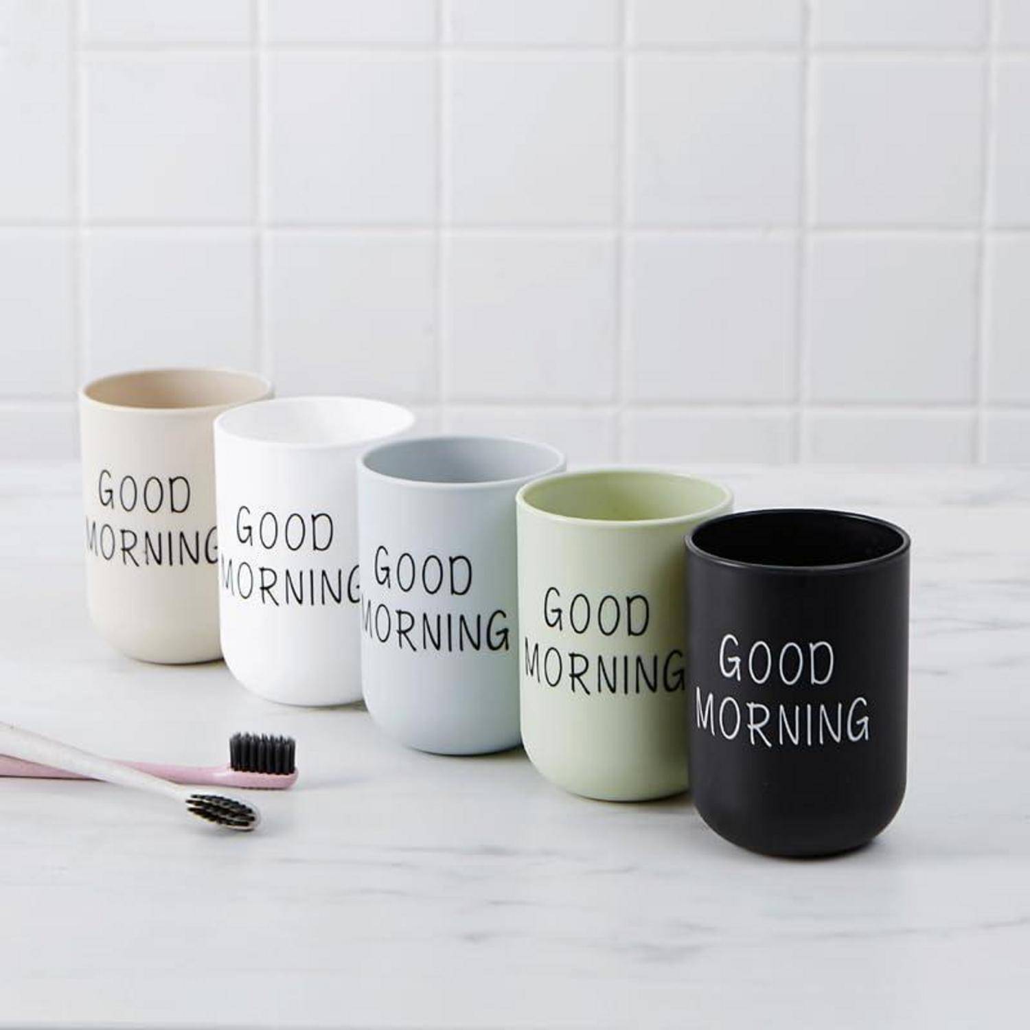 Fünf Keramiktassen in Creme, Weiß, Grau, Grün und Schwarz stehen nebeneinander auf einer Arbeitsplatte, jede bedruckt mit „GOOD MORNING