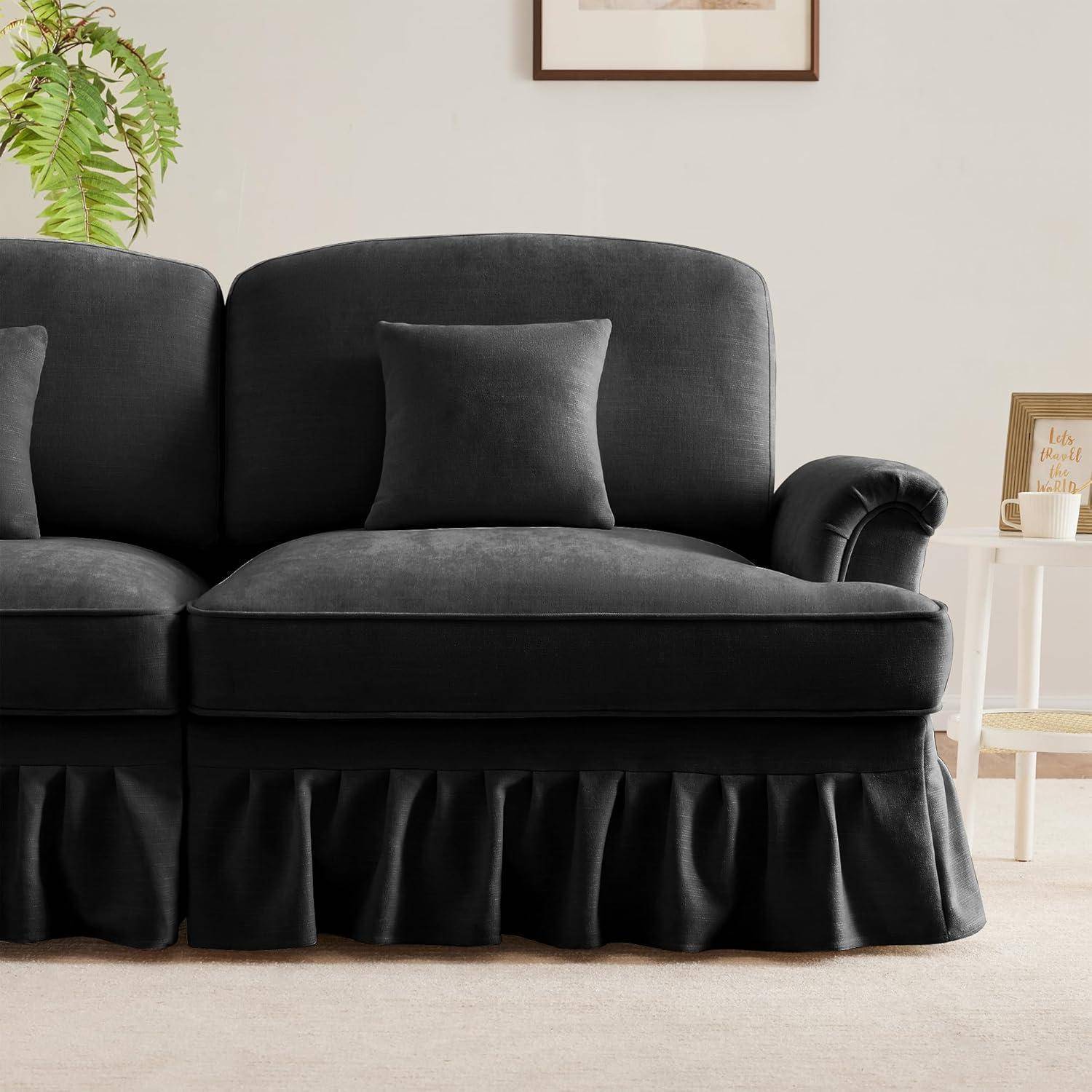 Schwarzes Polstersofa mit passendem quadratischem Kissen und Volantabdeckung in einem hellen Wohnzimmer, neben einem kleinen Beistelltisch und Wandkunst.