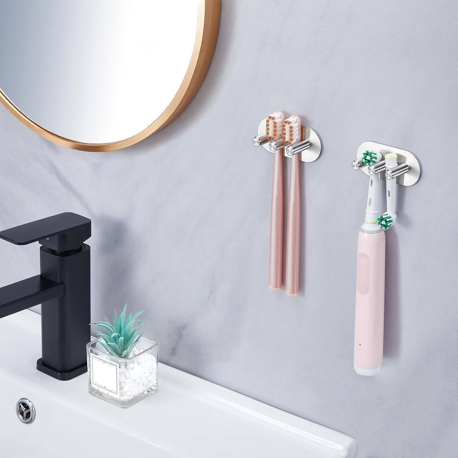 Wandmontierte Zahnbürstenhalter mit zwei Zahnbürsten und einer rosa elektrischen Zahnbürste neben einem Badezimmerspüle und Wasserhahn.