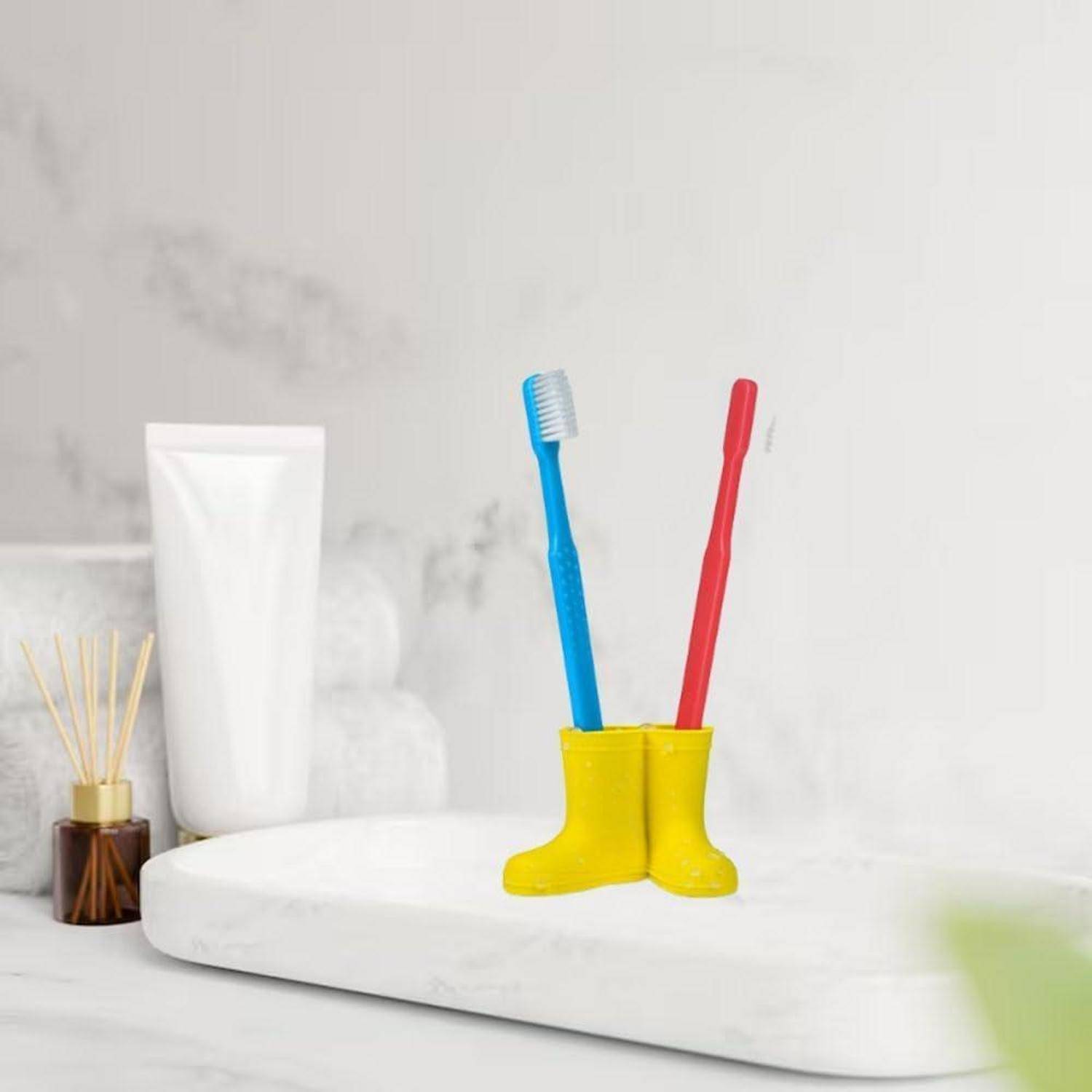 Zahnbürsten in einem gelben stiefelförmigen Behälter neben einer weißen Zahnpastatube, mit einem Raumduft-Diffuser auf einem weißen Badezimmerregal.