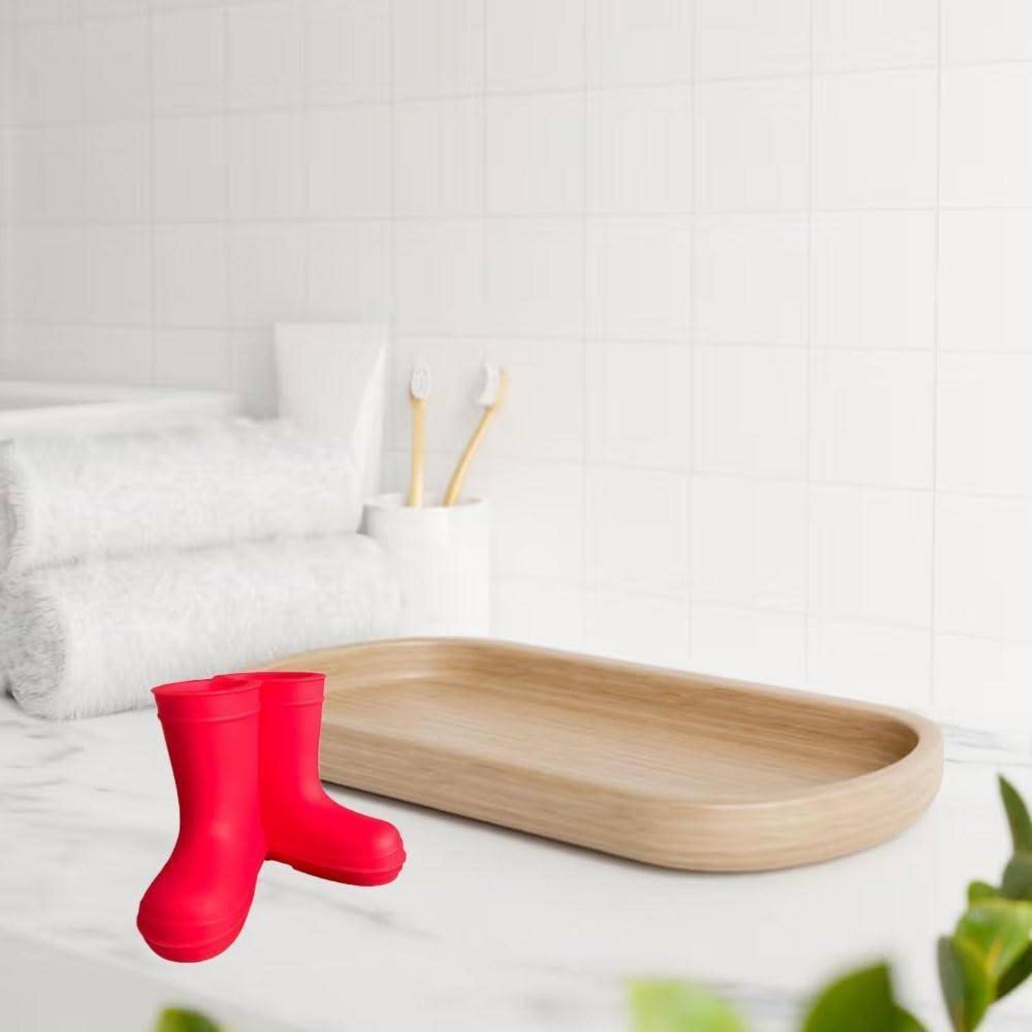 Rote Regenstiefel neben einem Holztablett in einem hellen Badezimmer oder einer Waschküche, mit Handtüchern und Zahnbürsten im Hintergrund.
